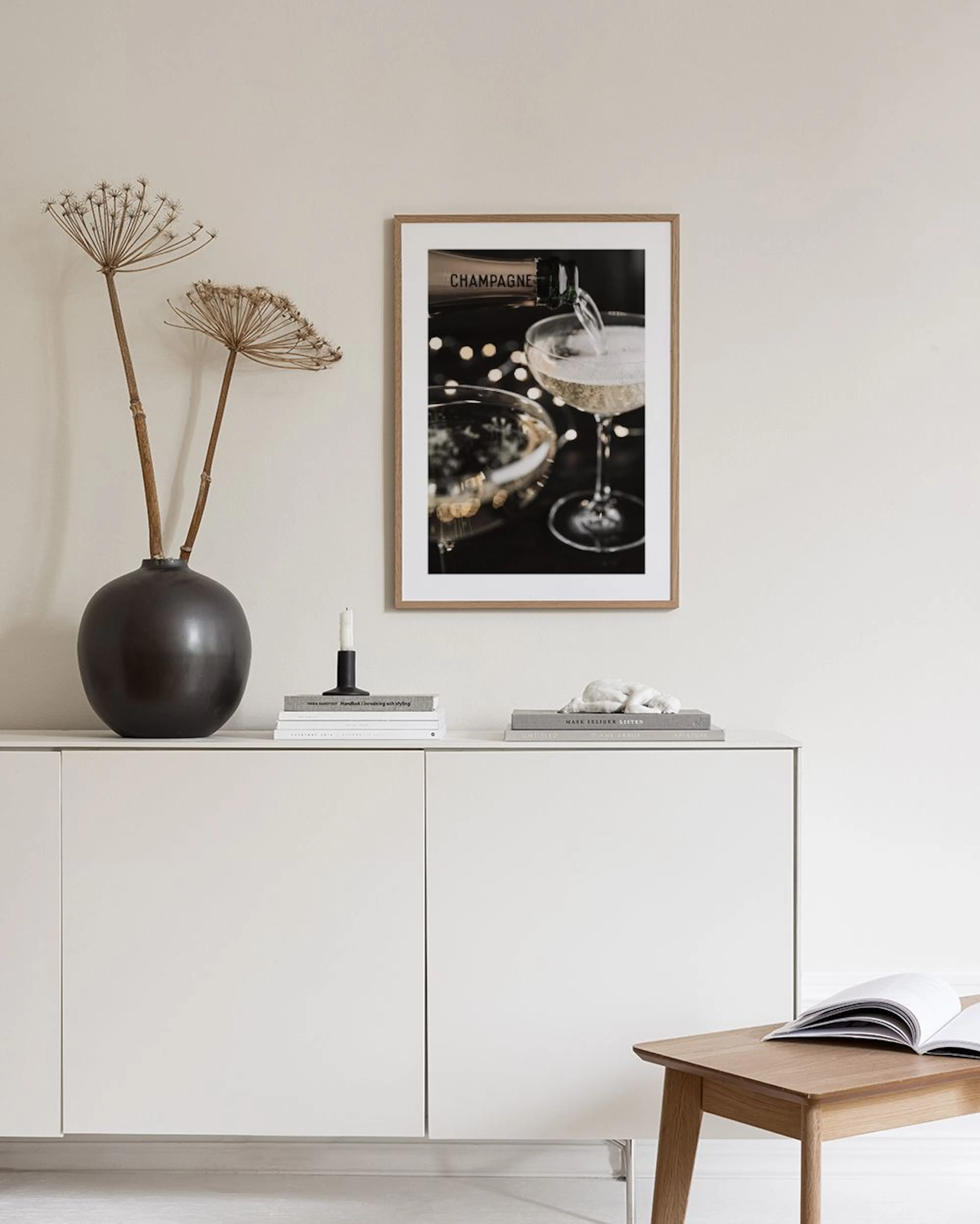 Plakat med champagne der hældes i glas, hængende på en grå væg over et hvidt bord med vaser og bøger.