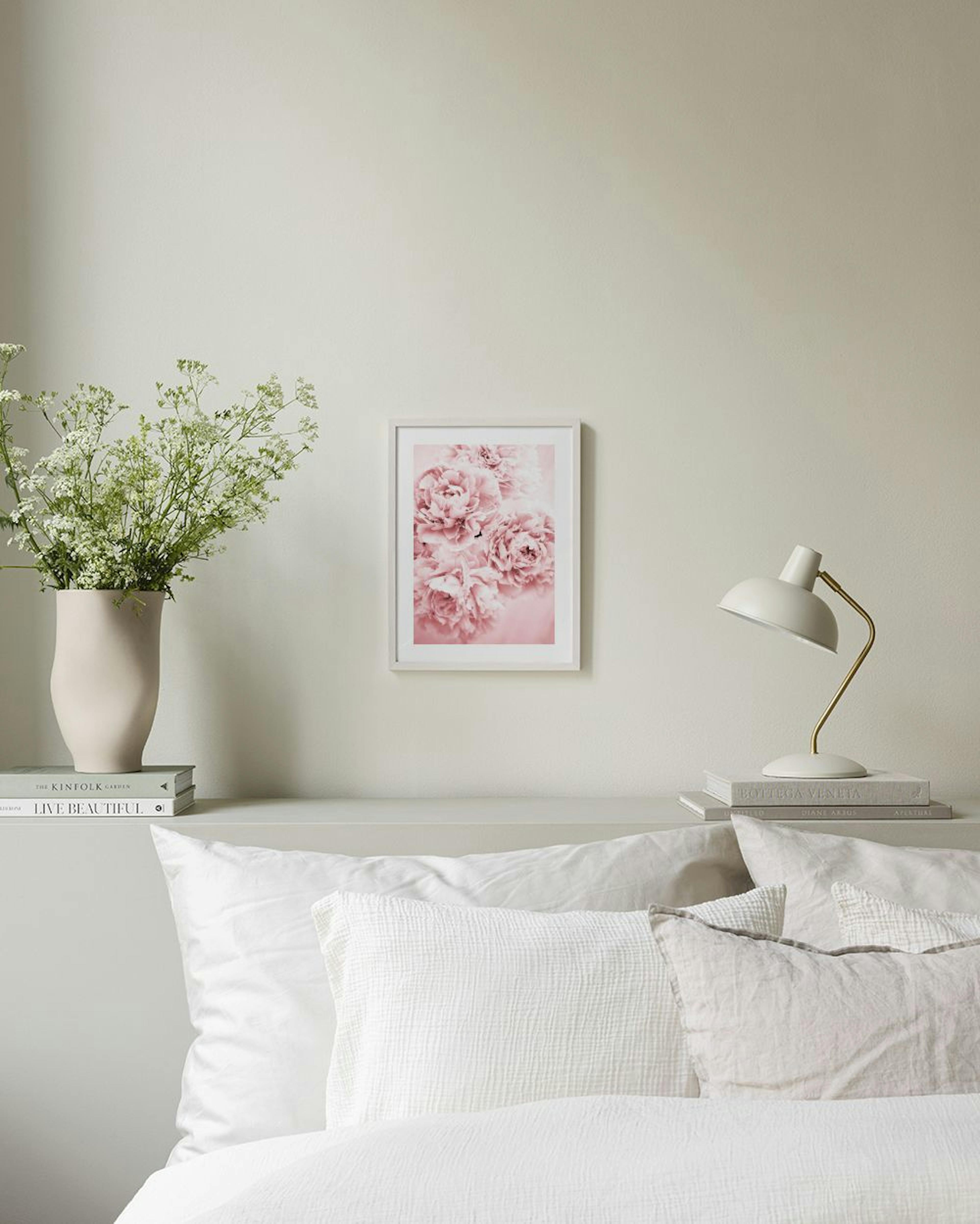 Poster med rosa pioner i en vit ram, hängande ovanför ett skrivbord med en stol, böcker och en vas.
