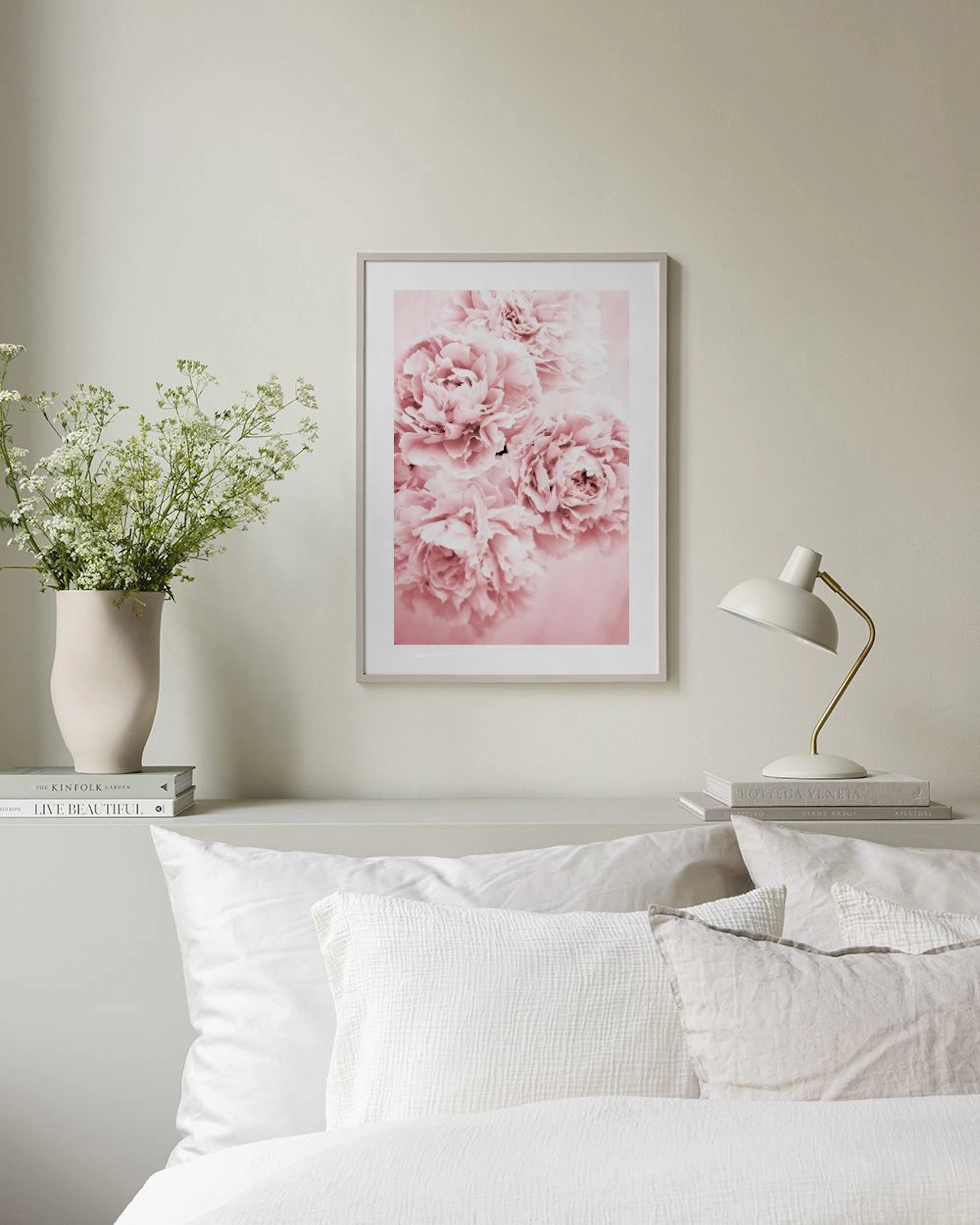 Poster med rosa pioner i en vit ram, hängande ovanför ett skrivbord med en stol, böcker och en vas.