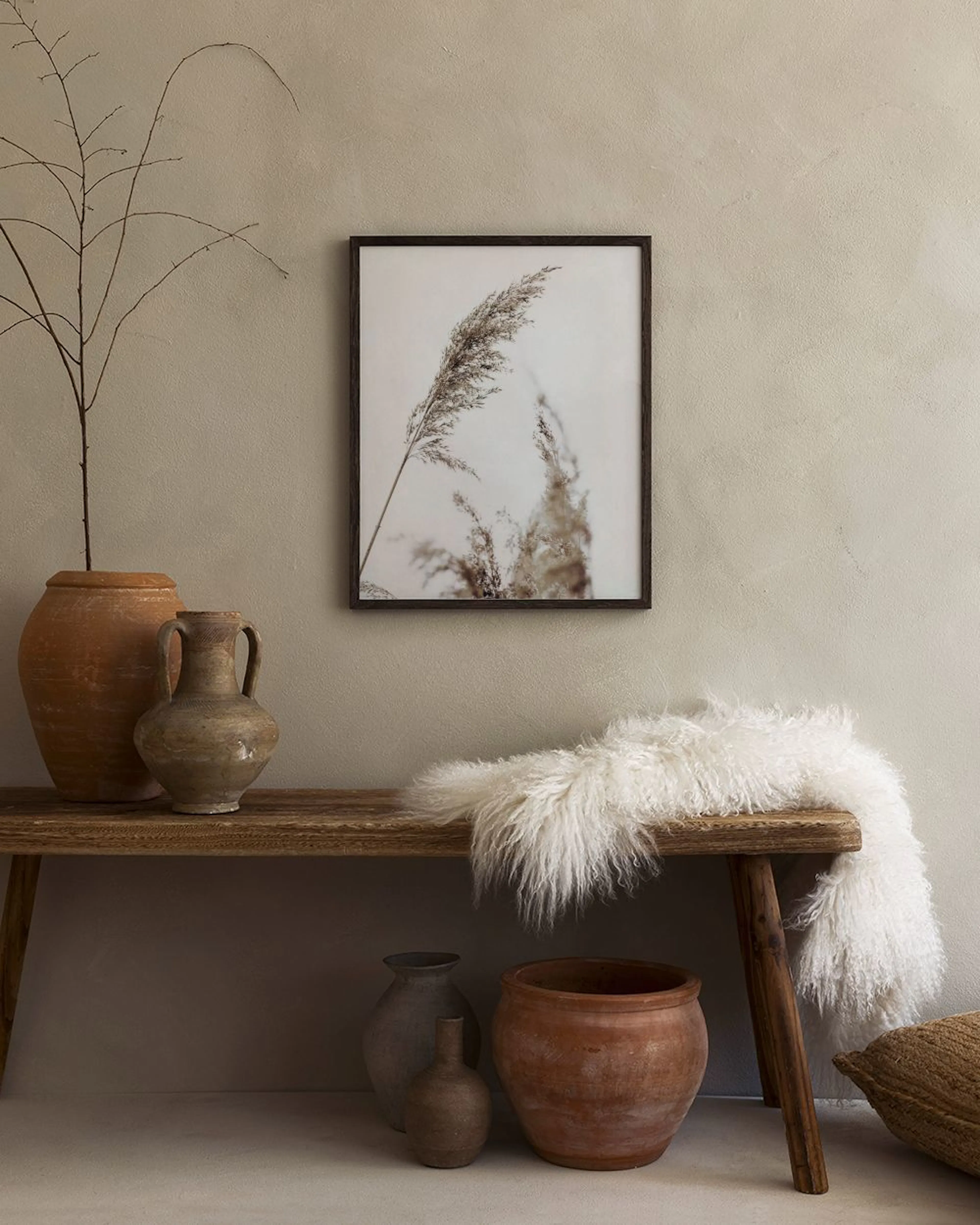 A print of dry pampas grass on a white background, hanging above a light wood desk with a chair and vase.