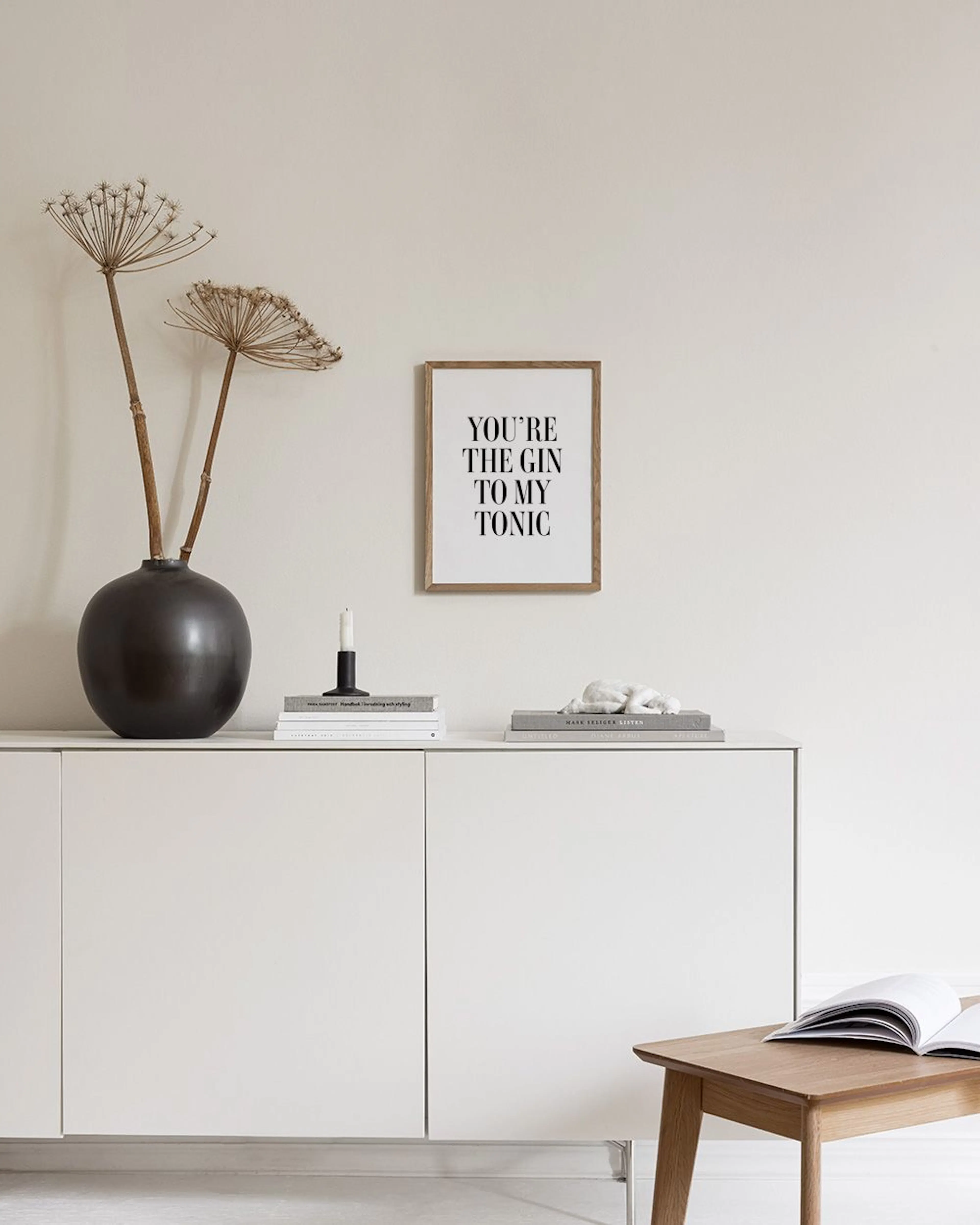 Um poster com a frase YOURE THE GIN TO MY TONIC em preto sobre fundo branco, exibido num móvel branco com um vaso e planta.