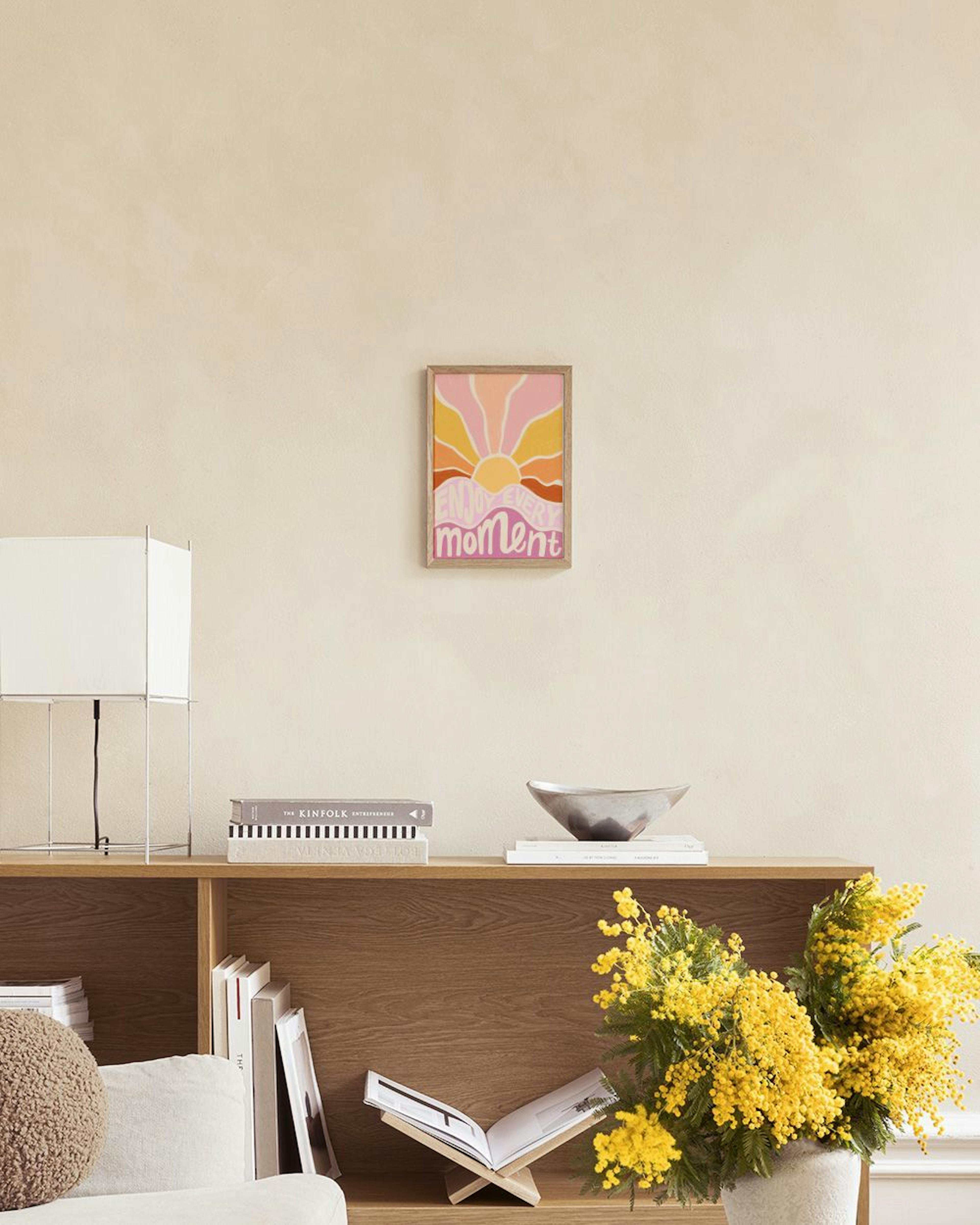 Poster mit Sonnenaufgang in Gelb, Orange und Rosa, Schriftzug Enjoy Every Moment“ in warmen Tönen, über einem Bett.