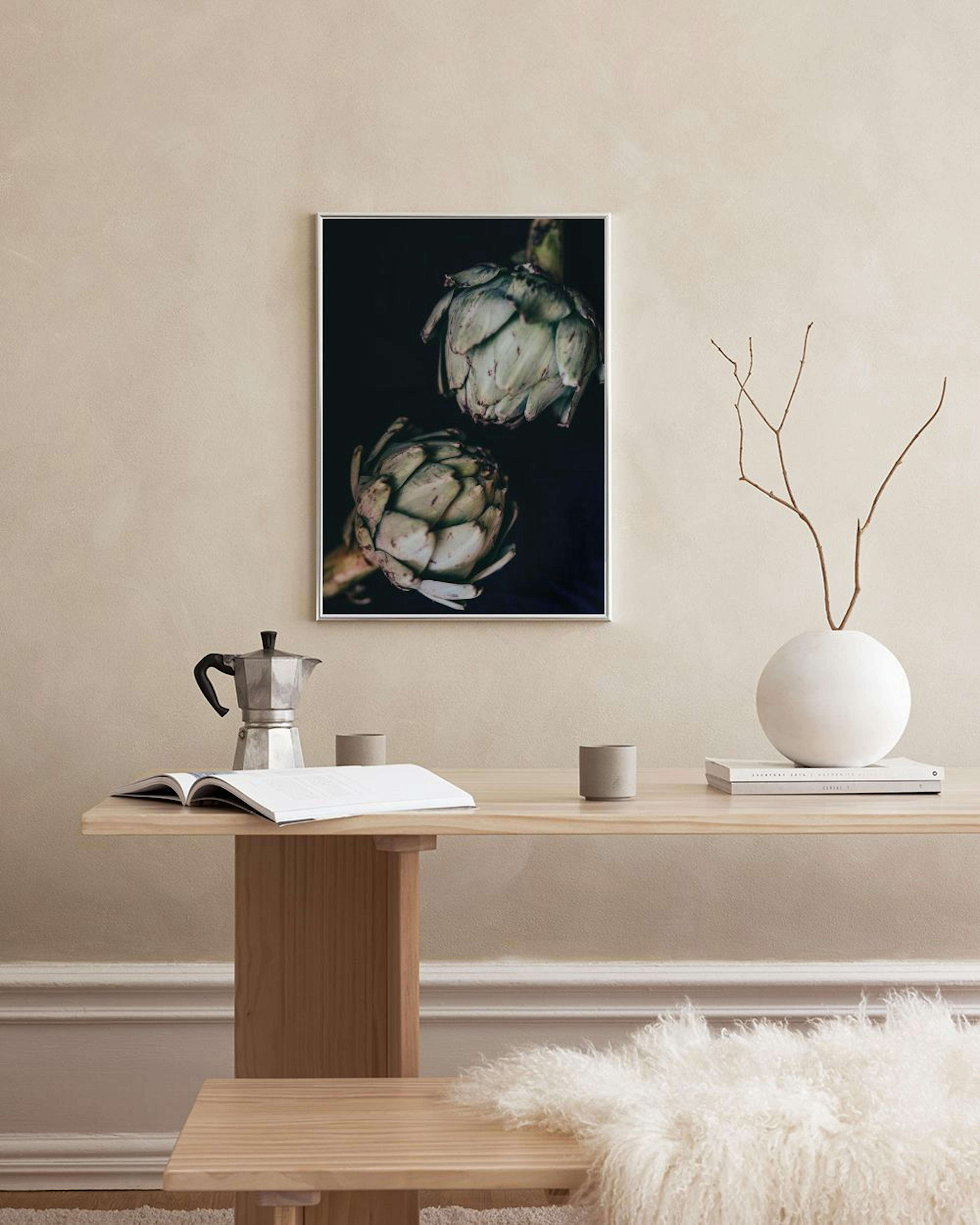 Póster oscuro con dos alcachofas colgando, exhibido en una pared blanca sobre una mesa con un mantel blanco.