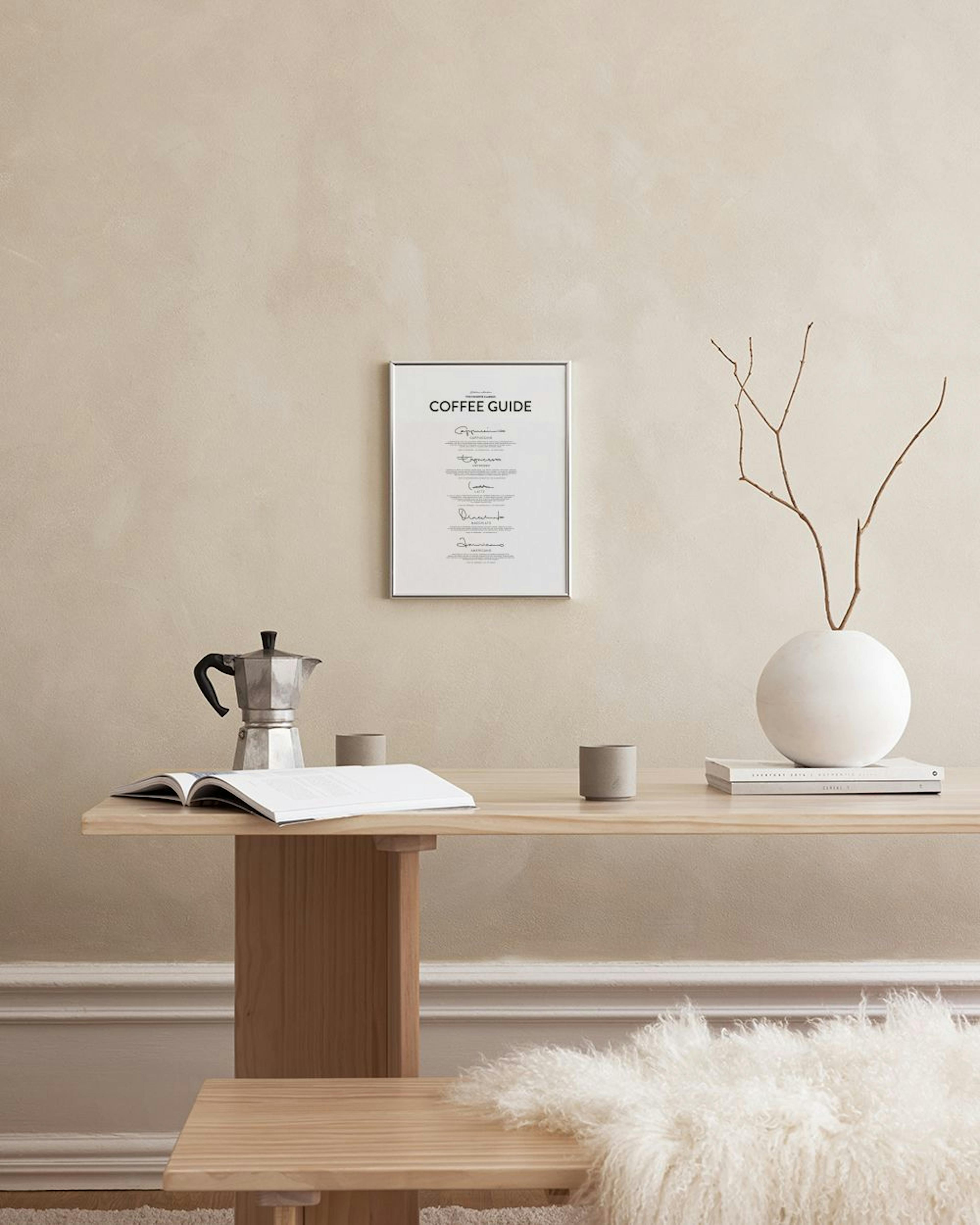 Biały plakat Coffee Guide ze stylizowanymi podpisami, ustawiony na białej półce na tle marmurowej ściany. Obok czarny wazon i no