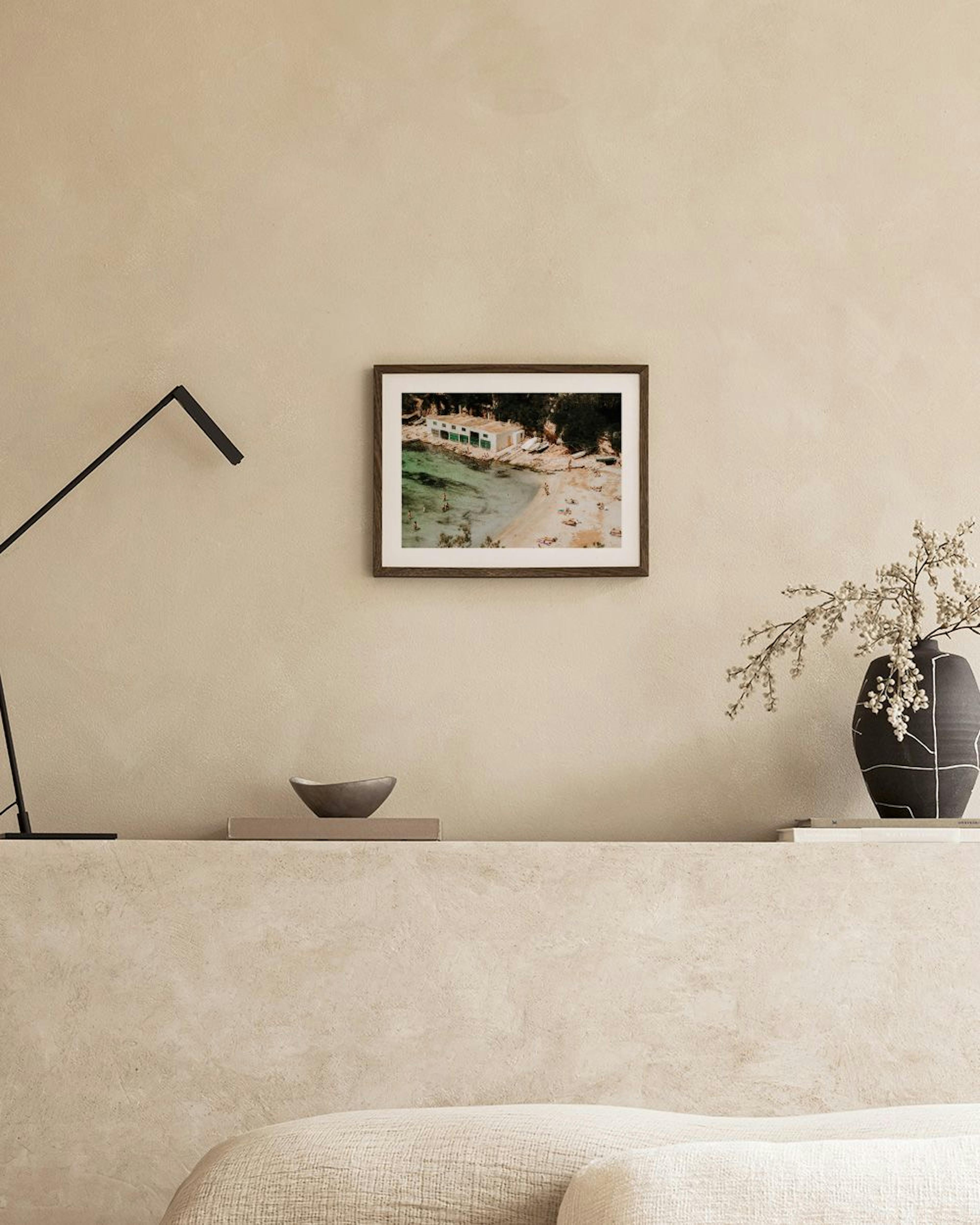 En poster med utsikt över en strand och ett hus vid havet, hängande ovanför en ljusbeige soffa.