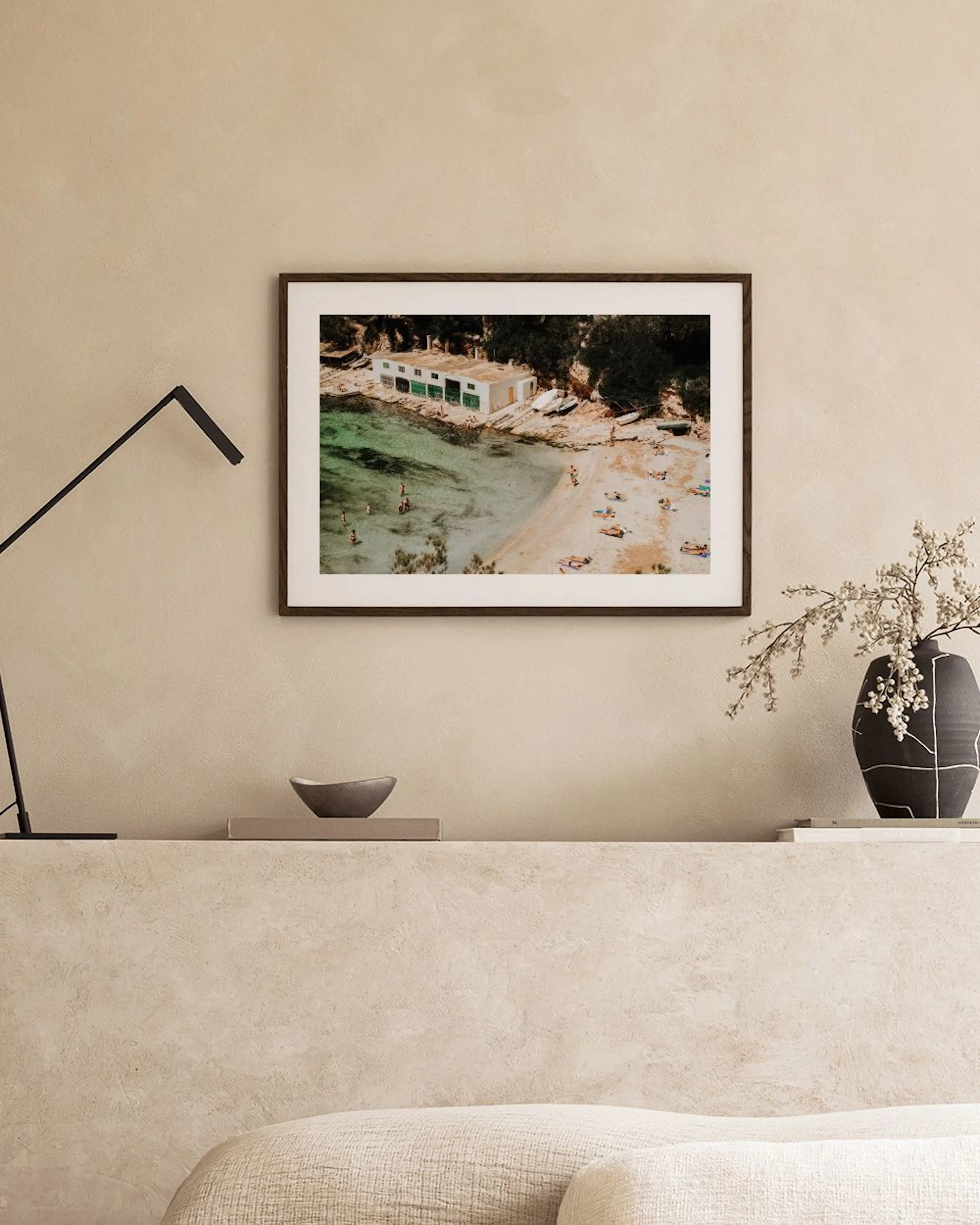 Affiche dune plage méditerranéenne avec des baigneurs et un cabanon blanc, suspendue au-dessus dun canapé beige.