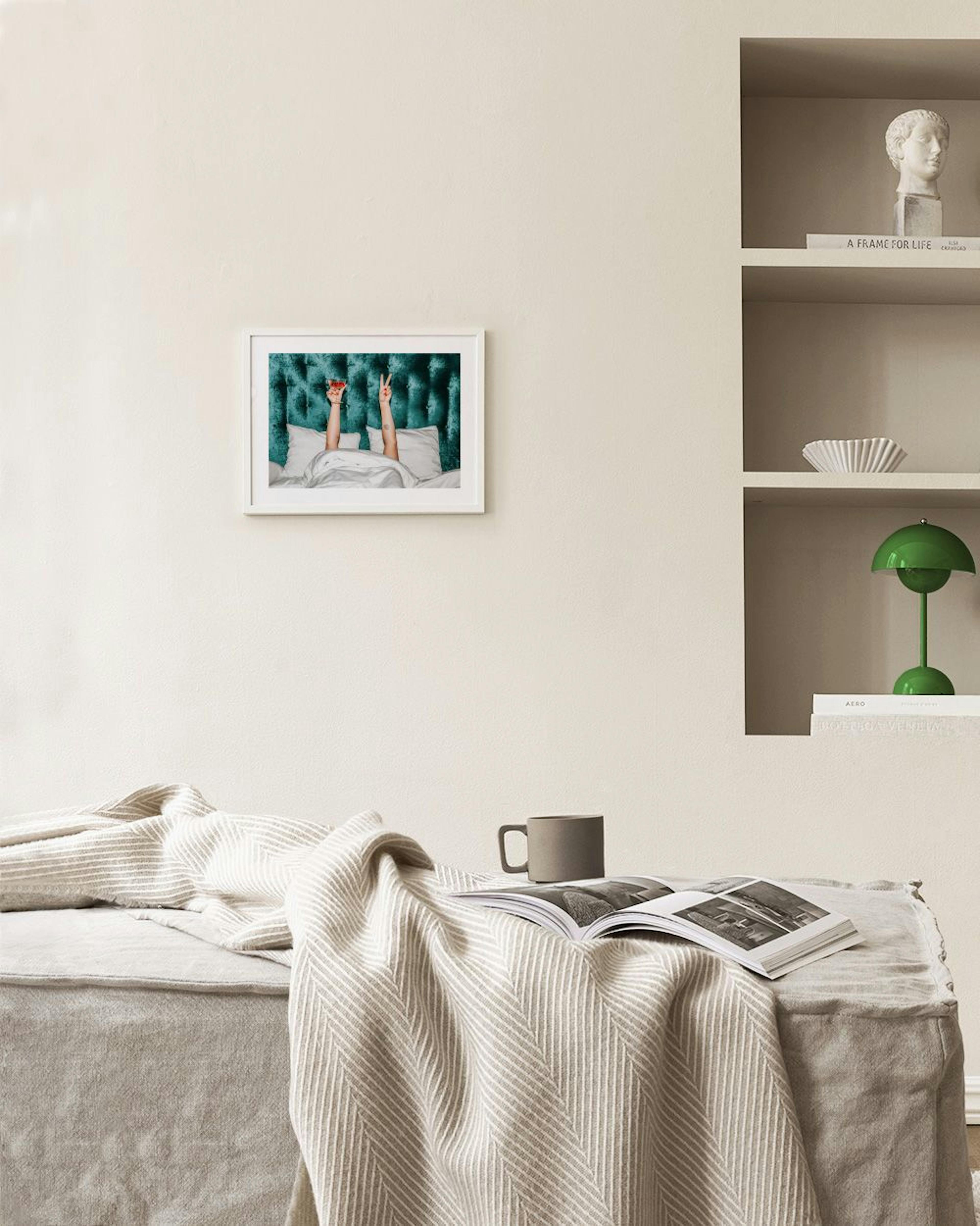 Póster de duas mãos a sair de um edredão branco com uma cama verde, exibido por cima de uma estante preta com livros e objetos d