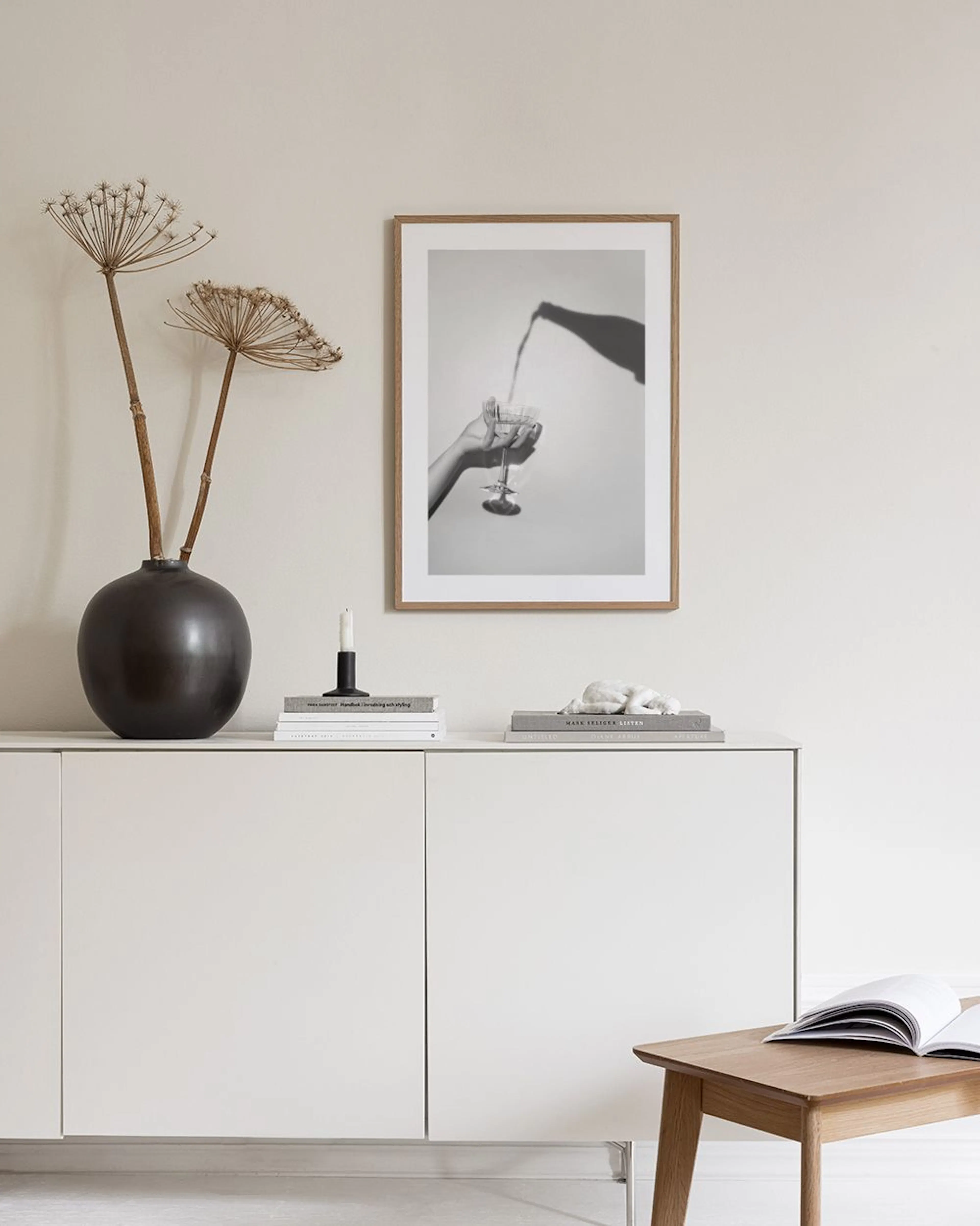 Poster in bianco e nero di una mano che tiene un bicchiere con ombra di bottiglia, su mobile bianco con vaso e libro.