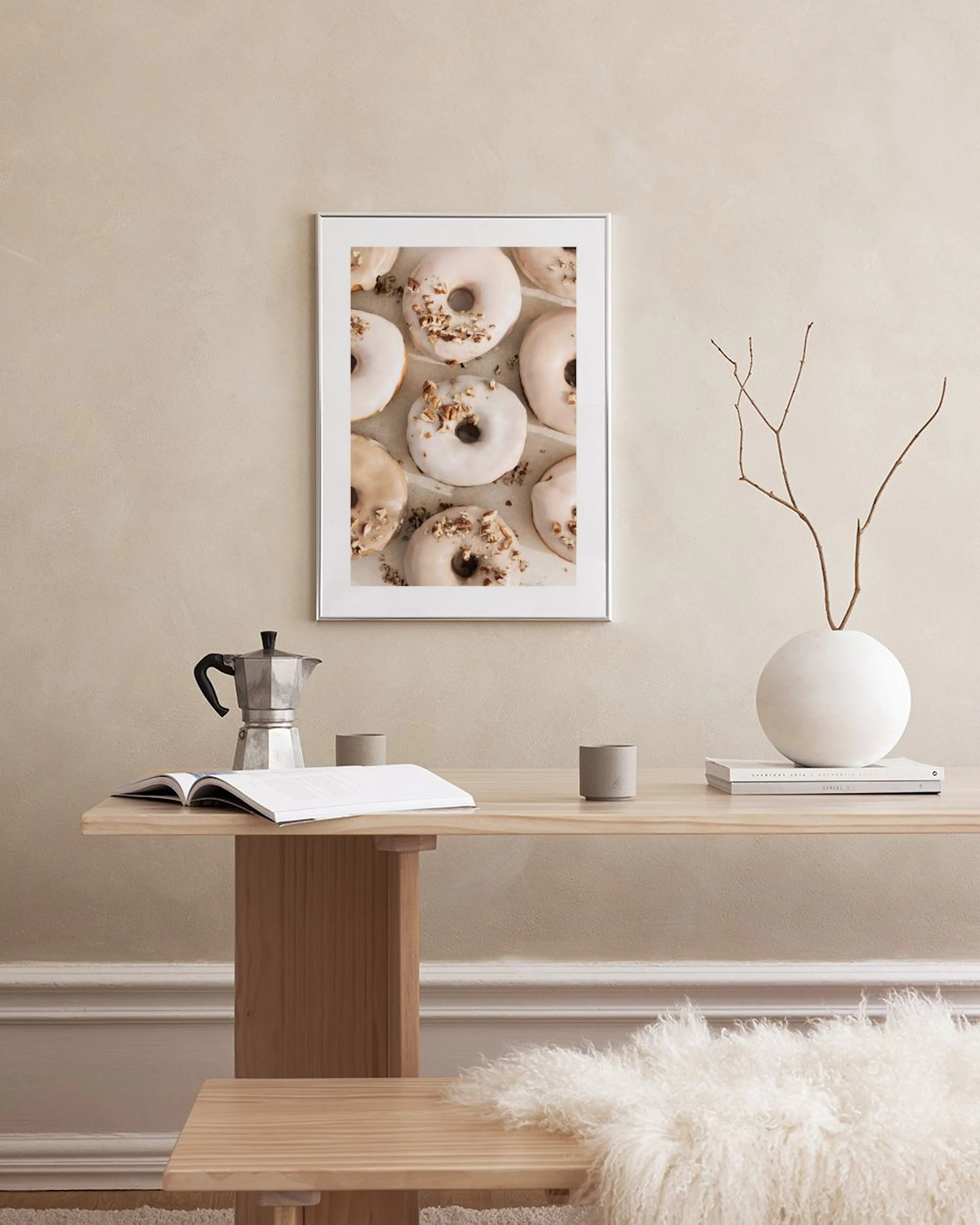 Plakat med lyse donuts med glasur og nødder, hængende over en sort hylde med en vase og bøger.