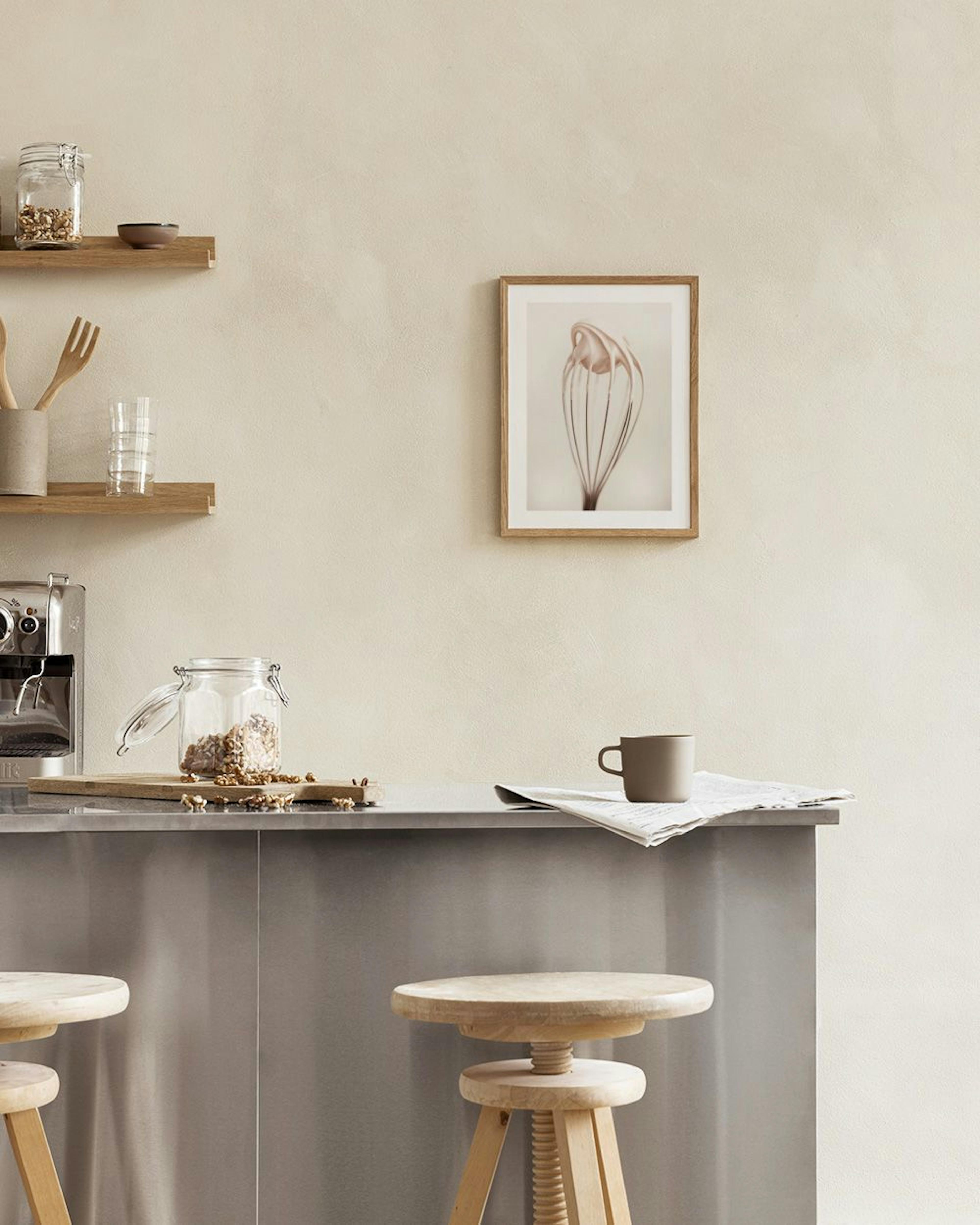 Póster de un batidor de globo con crema pastelera colgando en una cocina moderna sobre una mesa con un libro y una tetera.