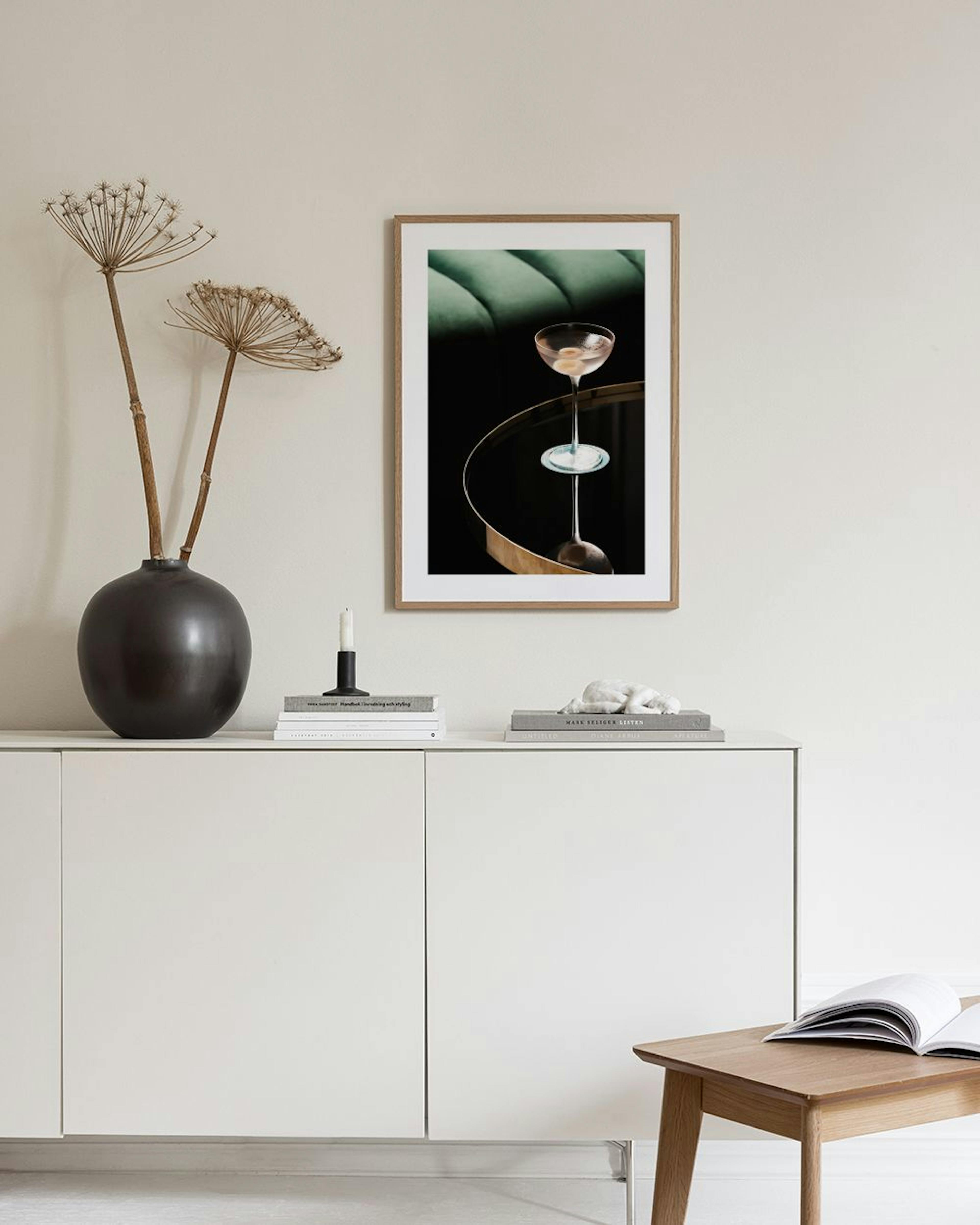 Poster med cocktailglas på runt bord, reflekterat i ytan. Hänger ovanför en vit byrå med vas och böcker.