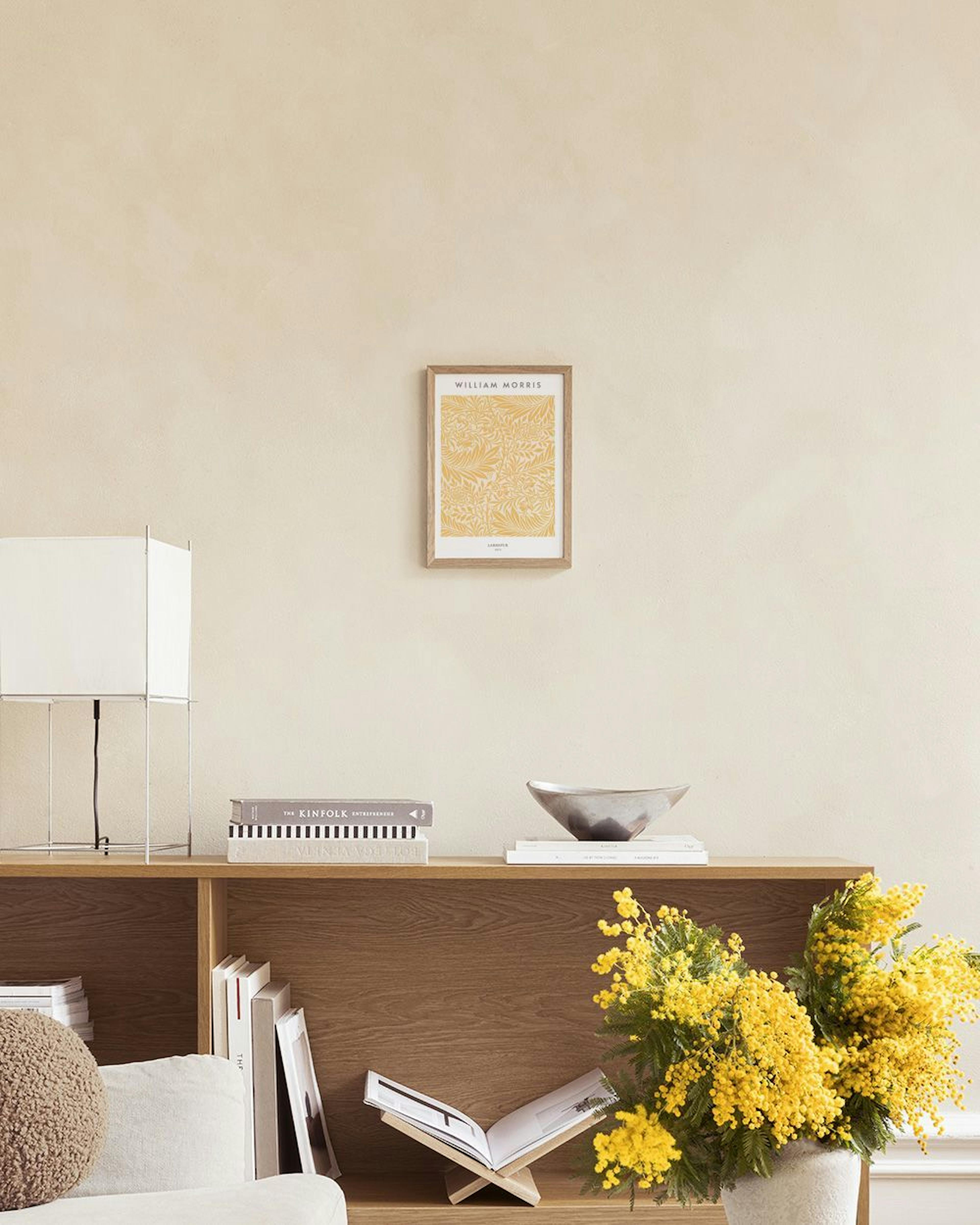 En William Morris-poster med ljust beiga blad och blommor, hängande ovanför ett sidobord med ljus och vas intill en vit fåtölj i