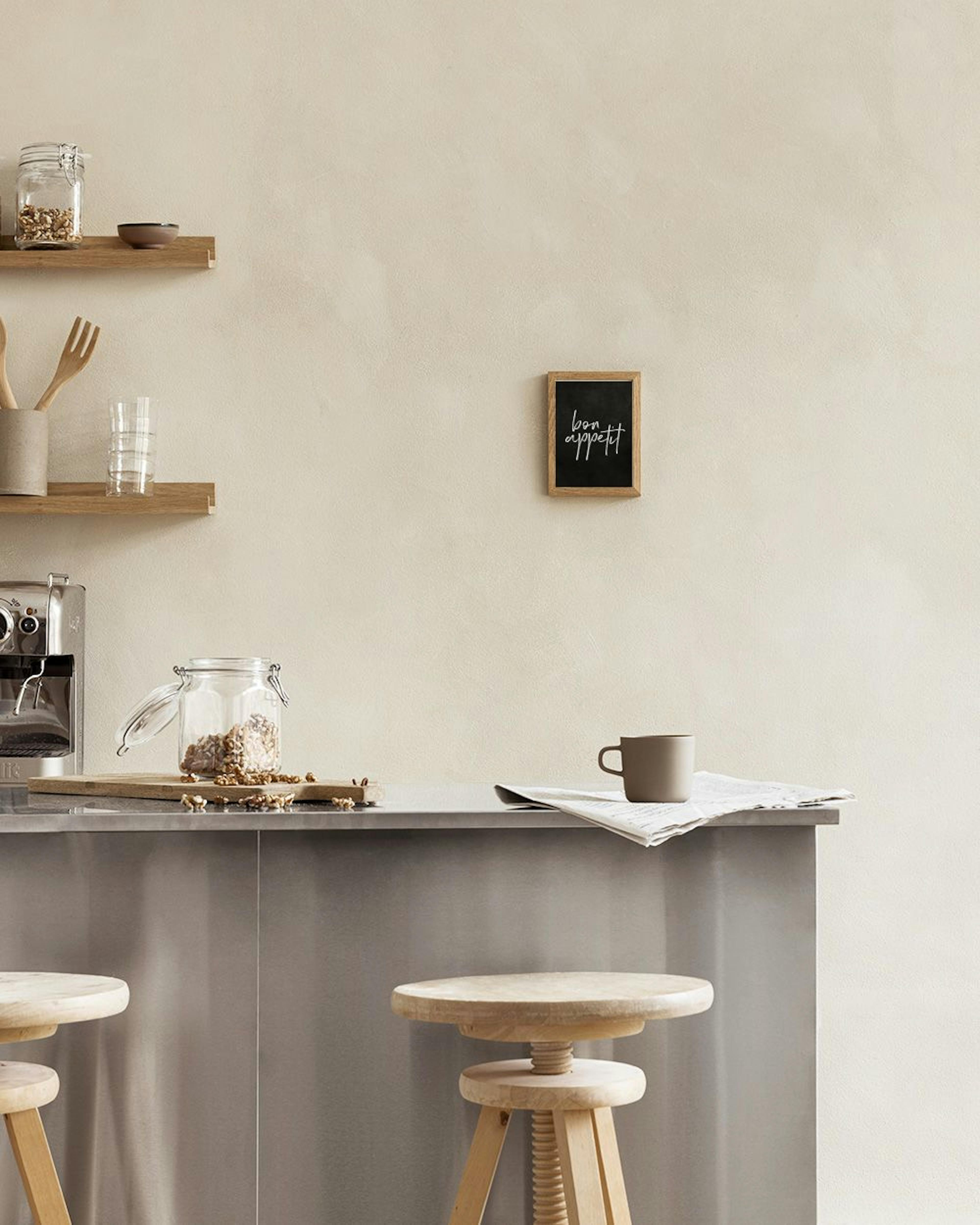 A print with bon appetit in white script on a black background, hanging above white drawers next to a glass bottle and fruit.