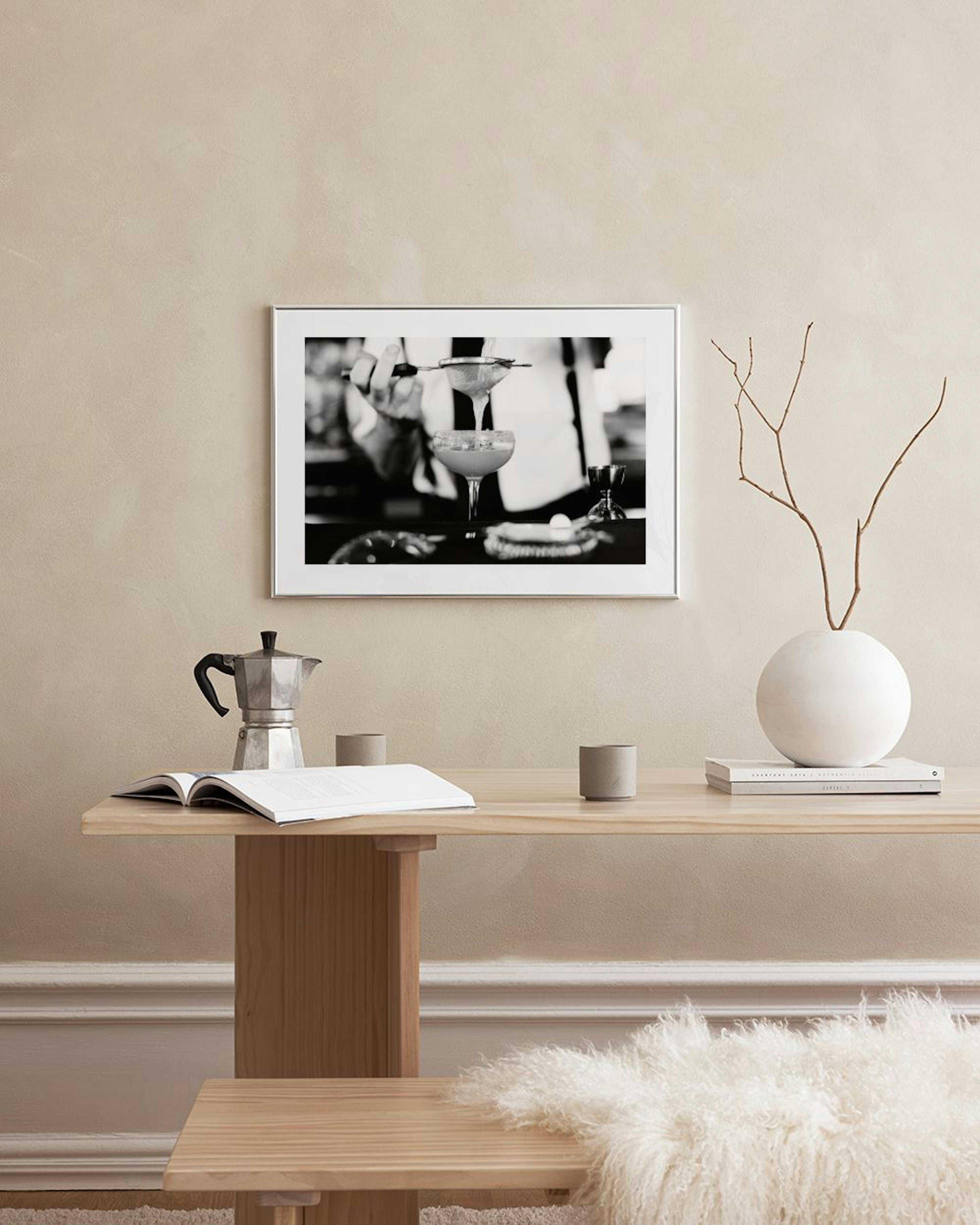A black and white print of a bartender pouring a drink, hanging on a light grey wall above a tufted ottoman.