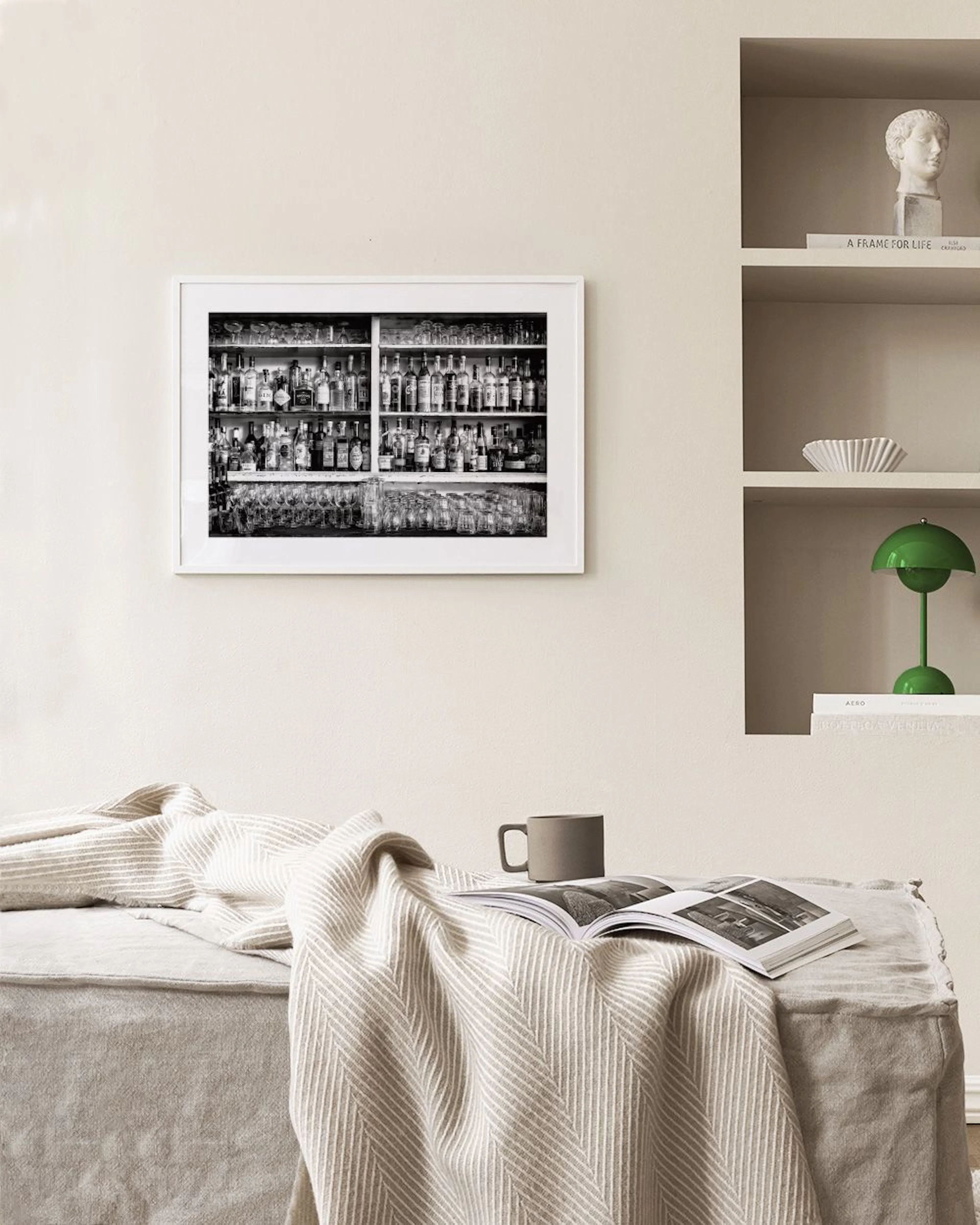 Black and white print of liquor bottles and glasses on a bar shelf, displayed above a gray buffet with books and a lamp.