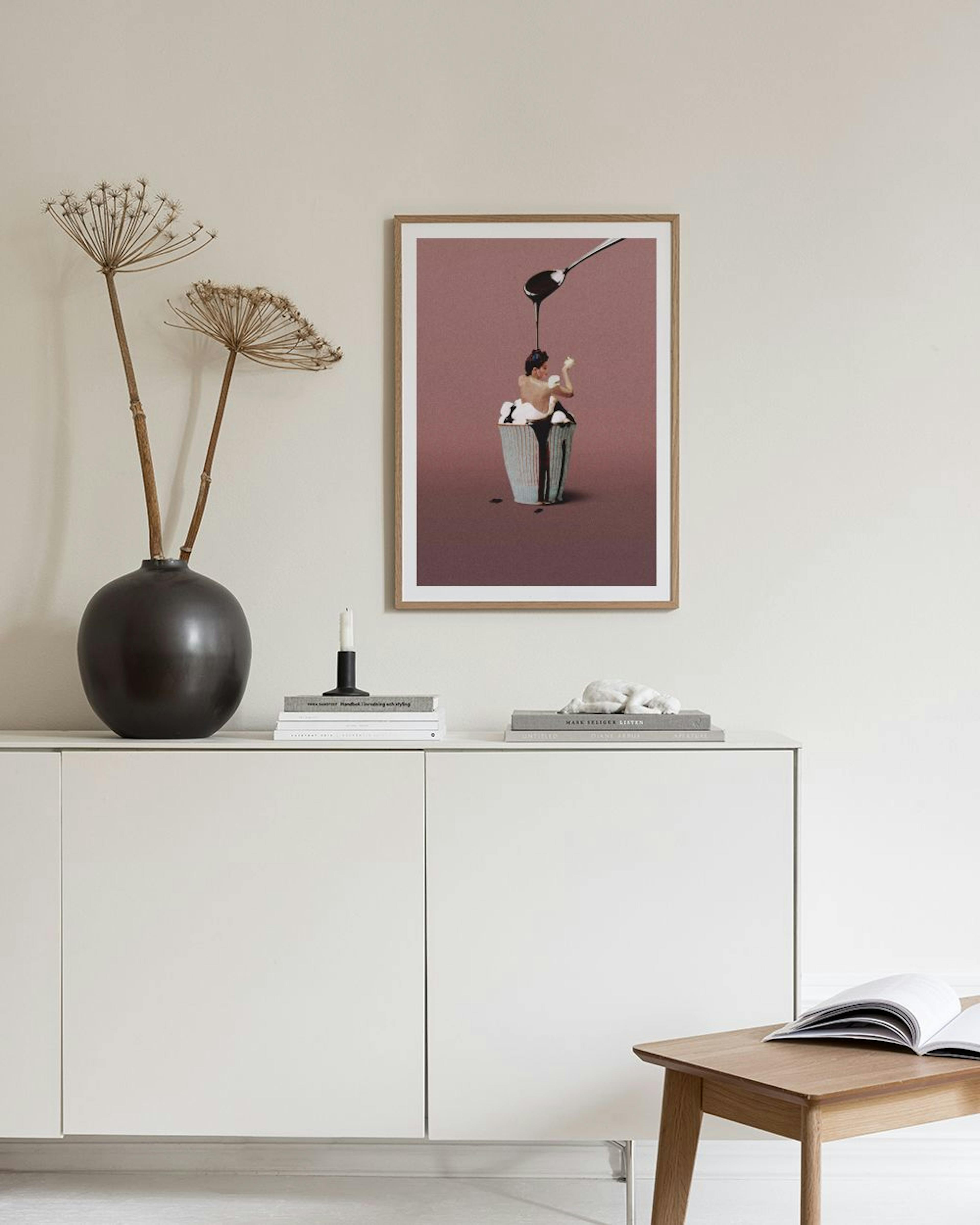 A print of a person in a dessert cup with melted chocolate pouring from above, hanging on a light gray wall.