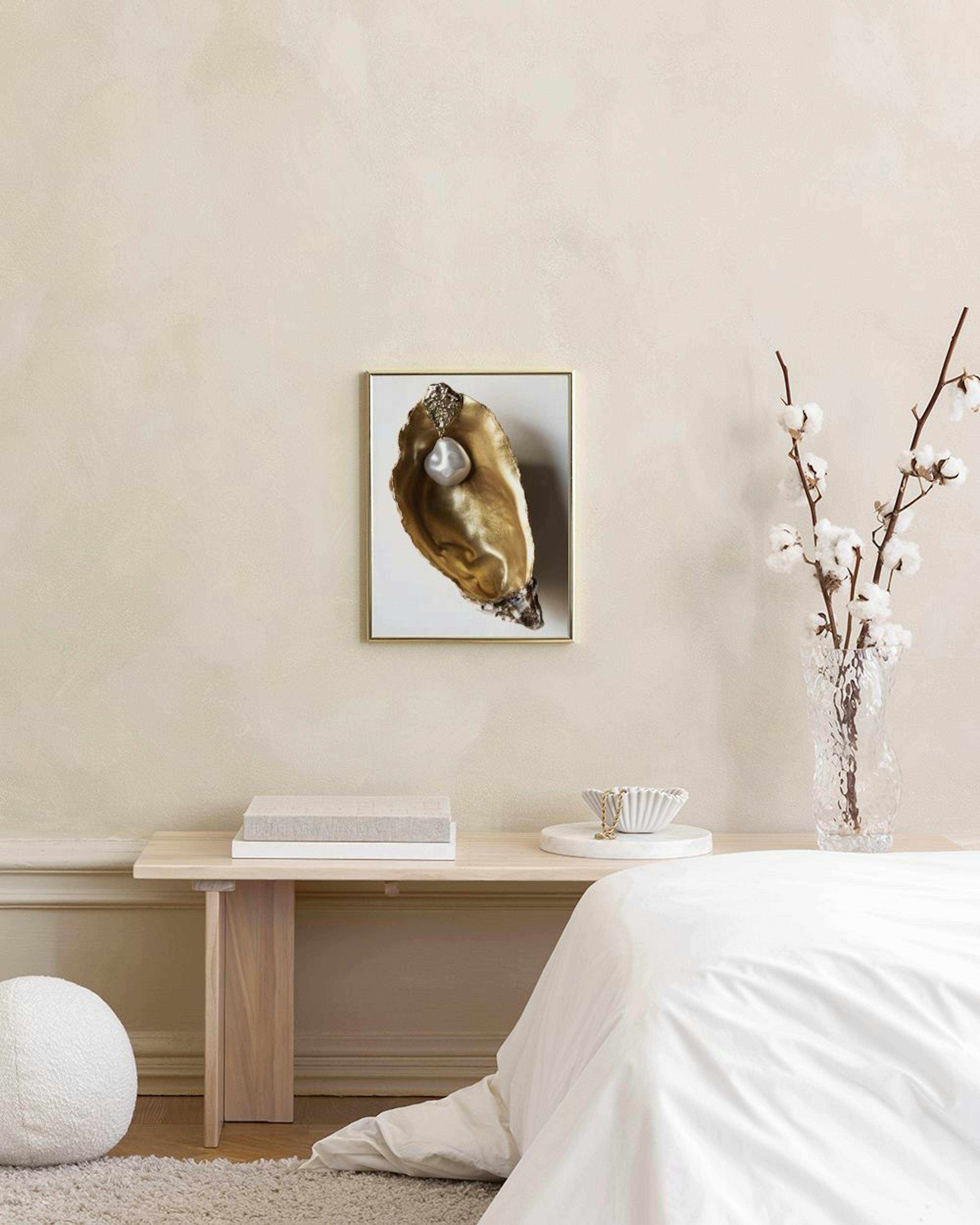 A print of a golden oyster shell with a large baroque pearl, displayed above a white console table with a black vase and stool.