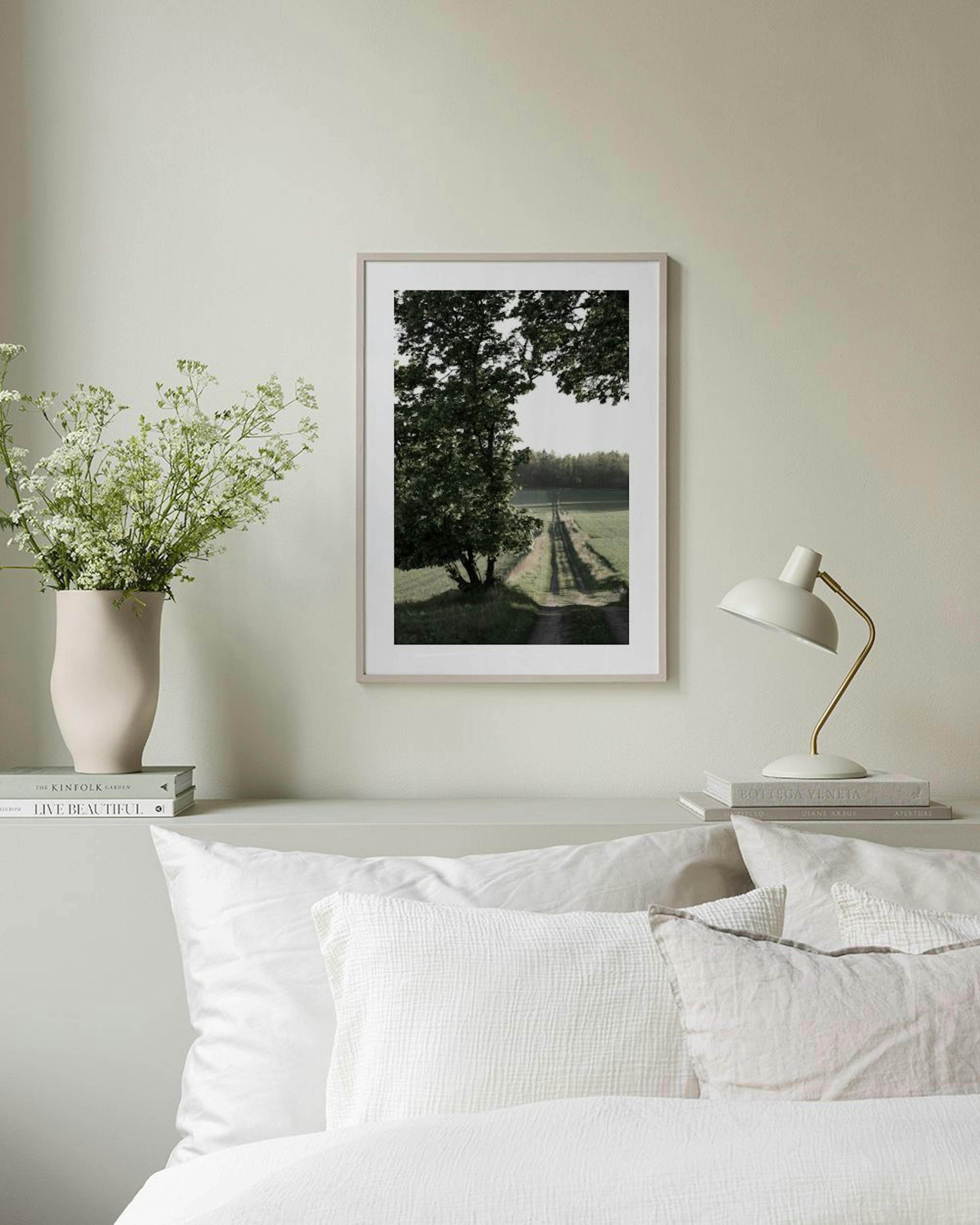 En poster med en grusväg bland gröna fält och träd, som hänger på en beige vägg ovanför ett mörkgrönt skåp.