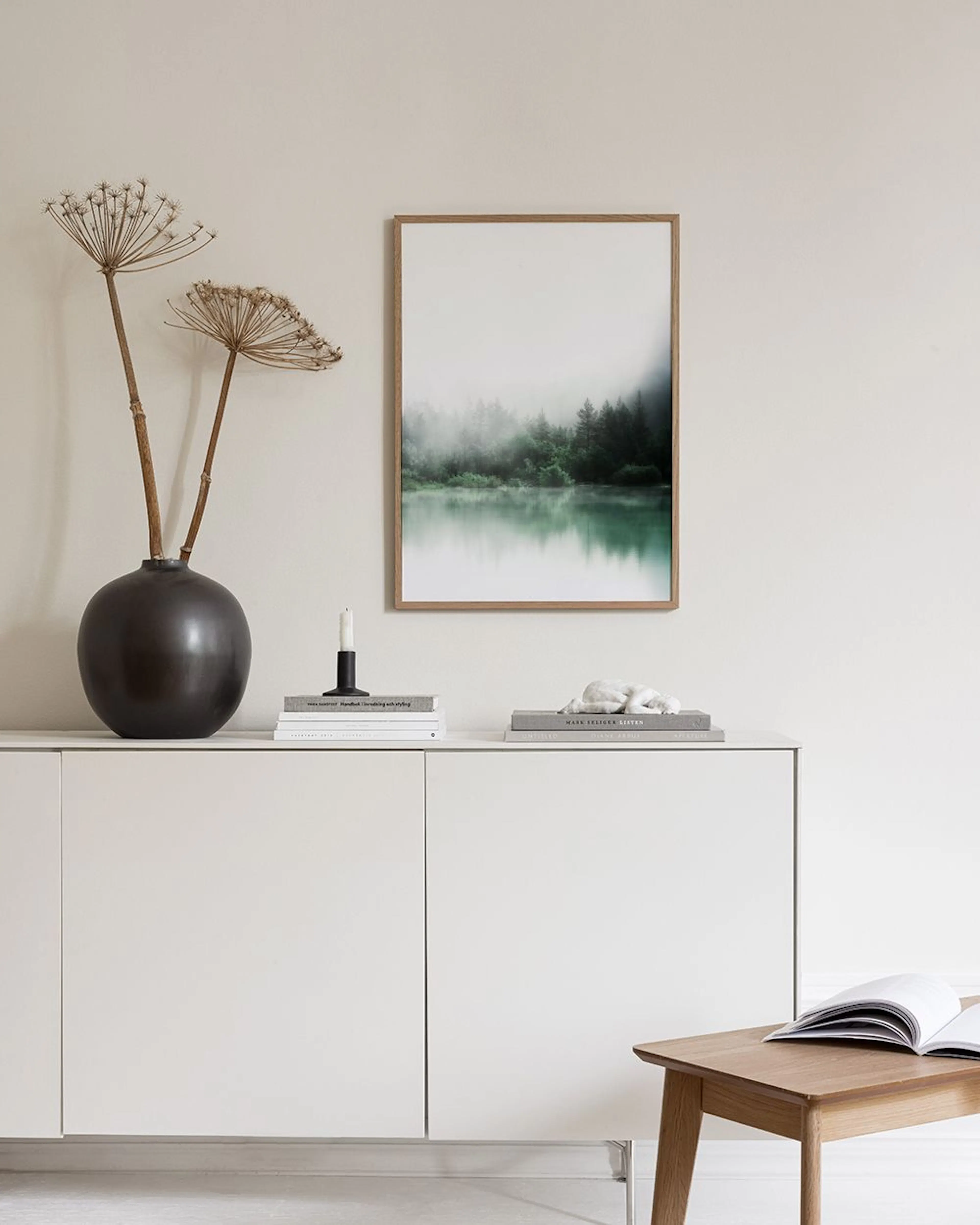 Affiche lac brumeux et forêt, suspendue au-dessus dun buffet blanc avec lampe et livres.