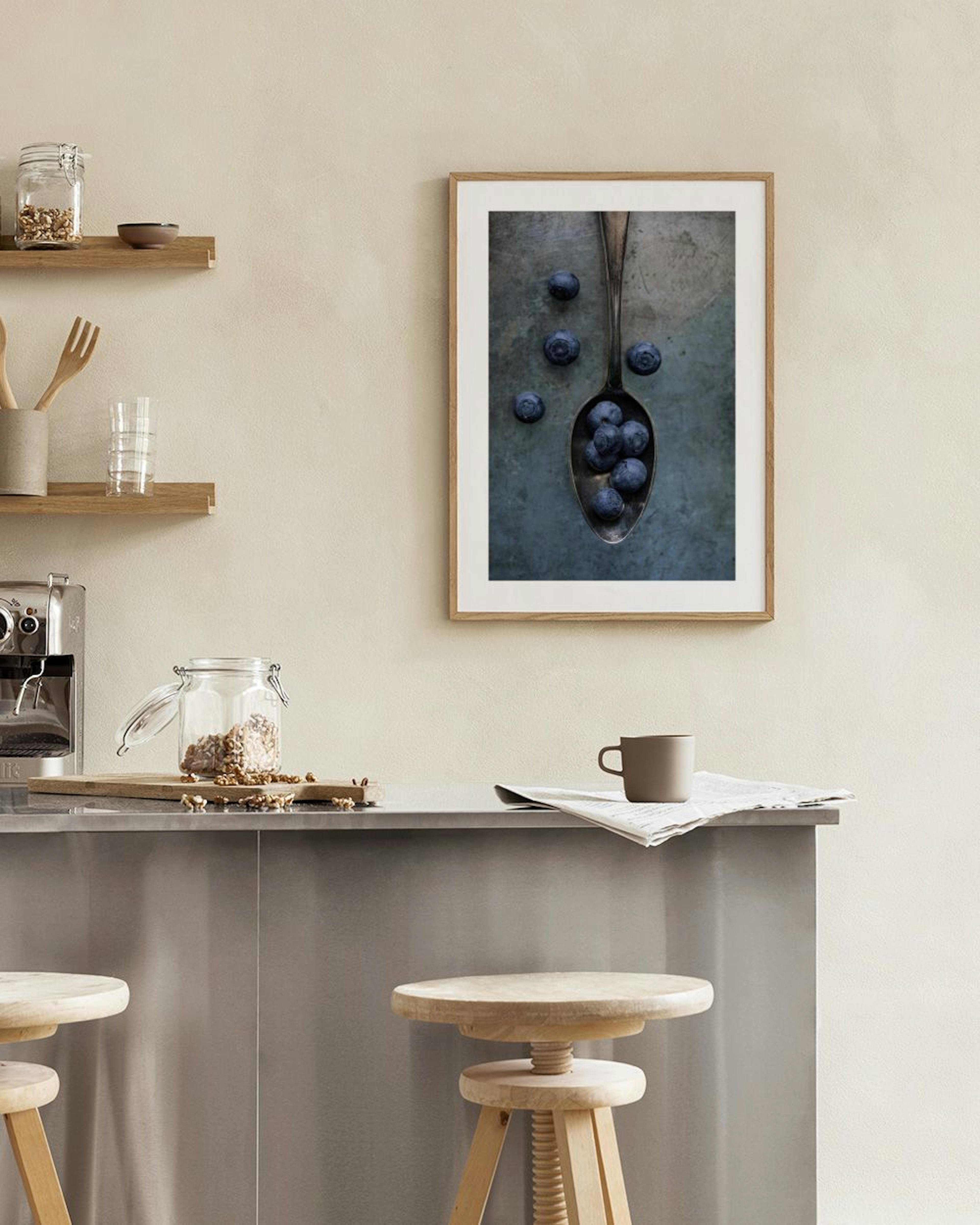 A print of blueberries in a spoon on a dark background, hanging on a white wall above a wooden chair and open book.
