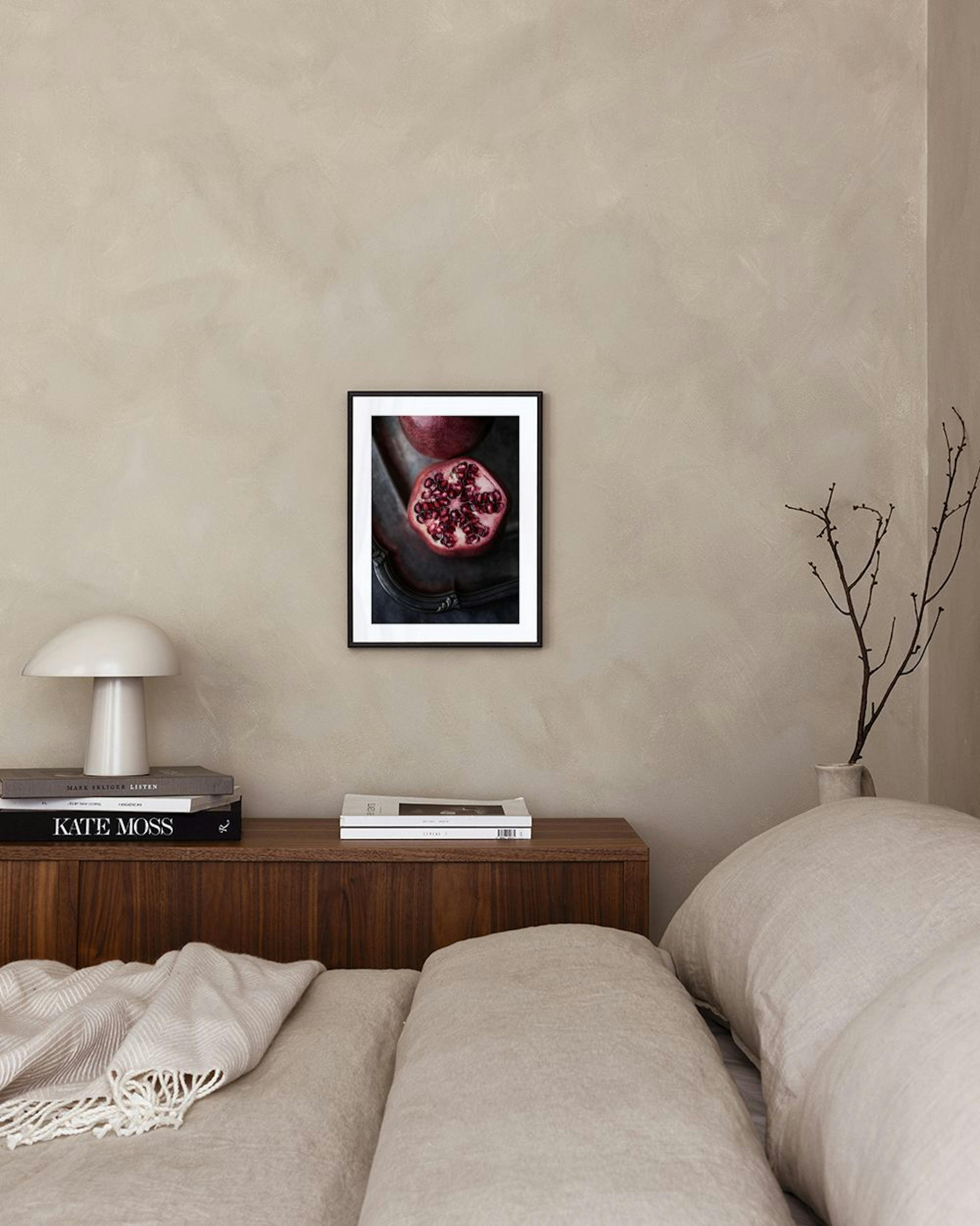 A print of a dark still life with a whole and cut pomegranate on a silver tray, hanging on a white wall above a wooden chair and