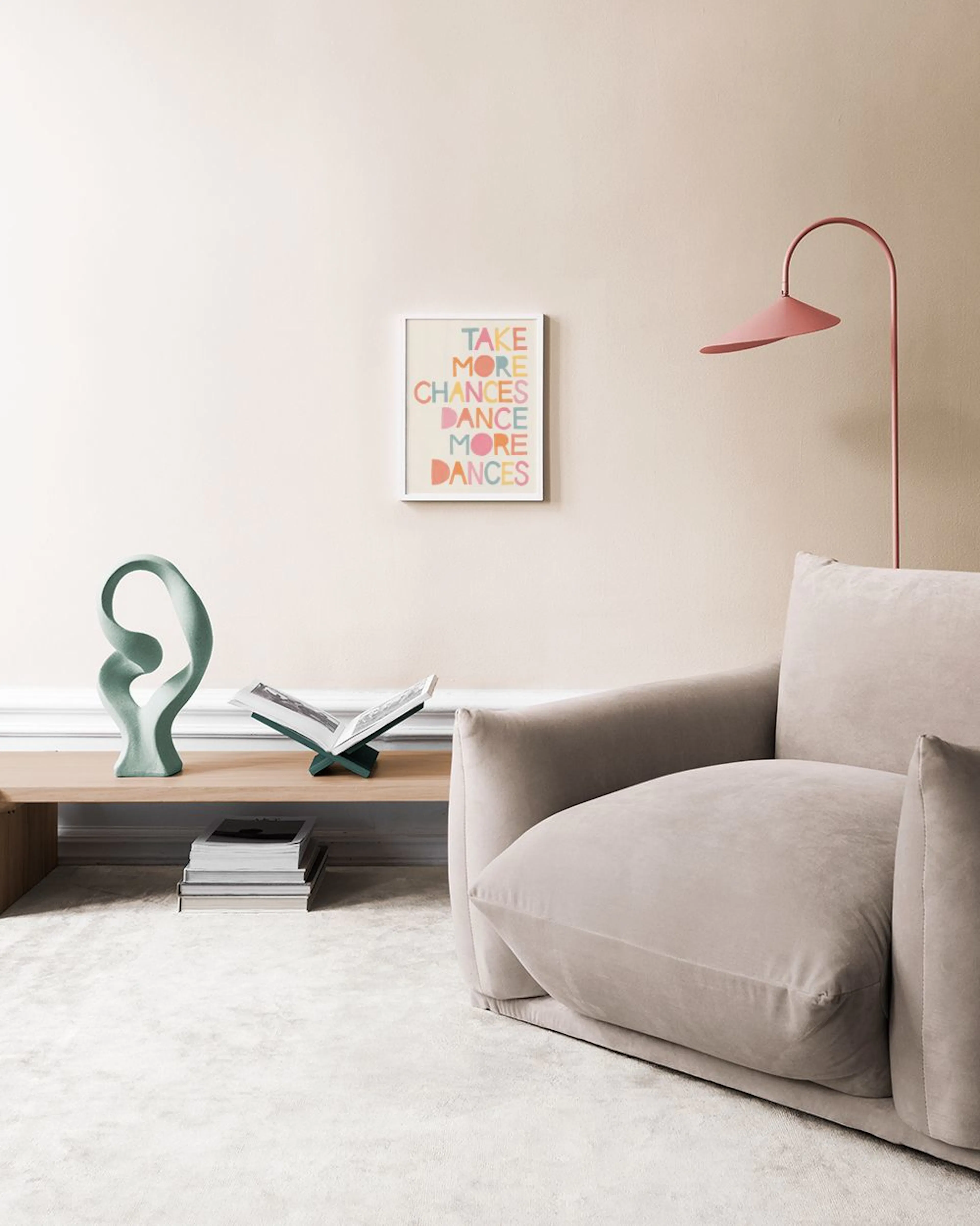 A colorful print with the text TAKE MORE CHANCES DANCE MORE DANCES in a room with a light gray vase and green plant.