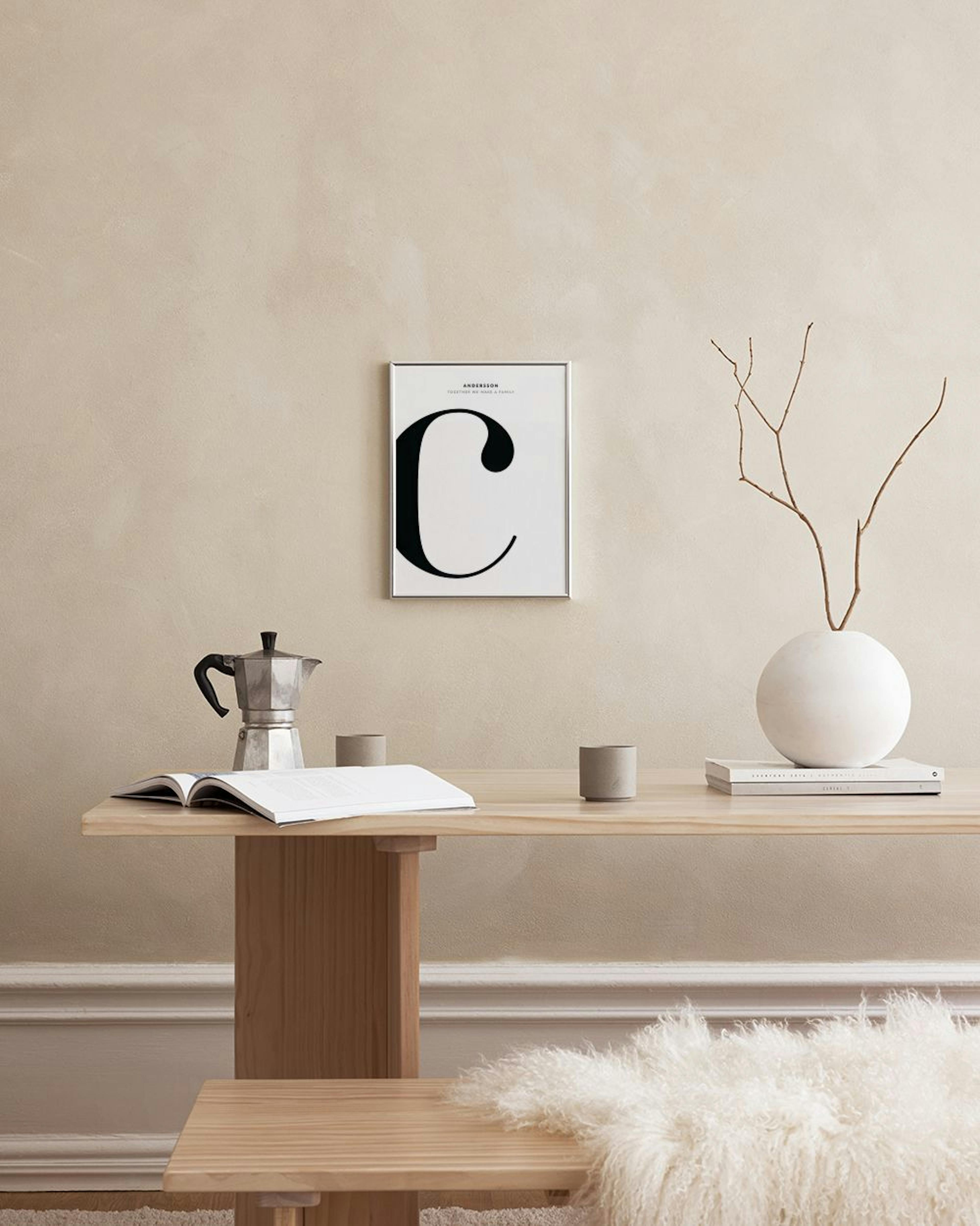 A personalized print with a large black letter C, ANDERSSON, and TOGETHER WE MAKE A FAMILY text, displayed above a light wood ca