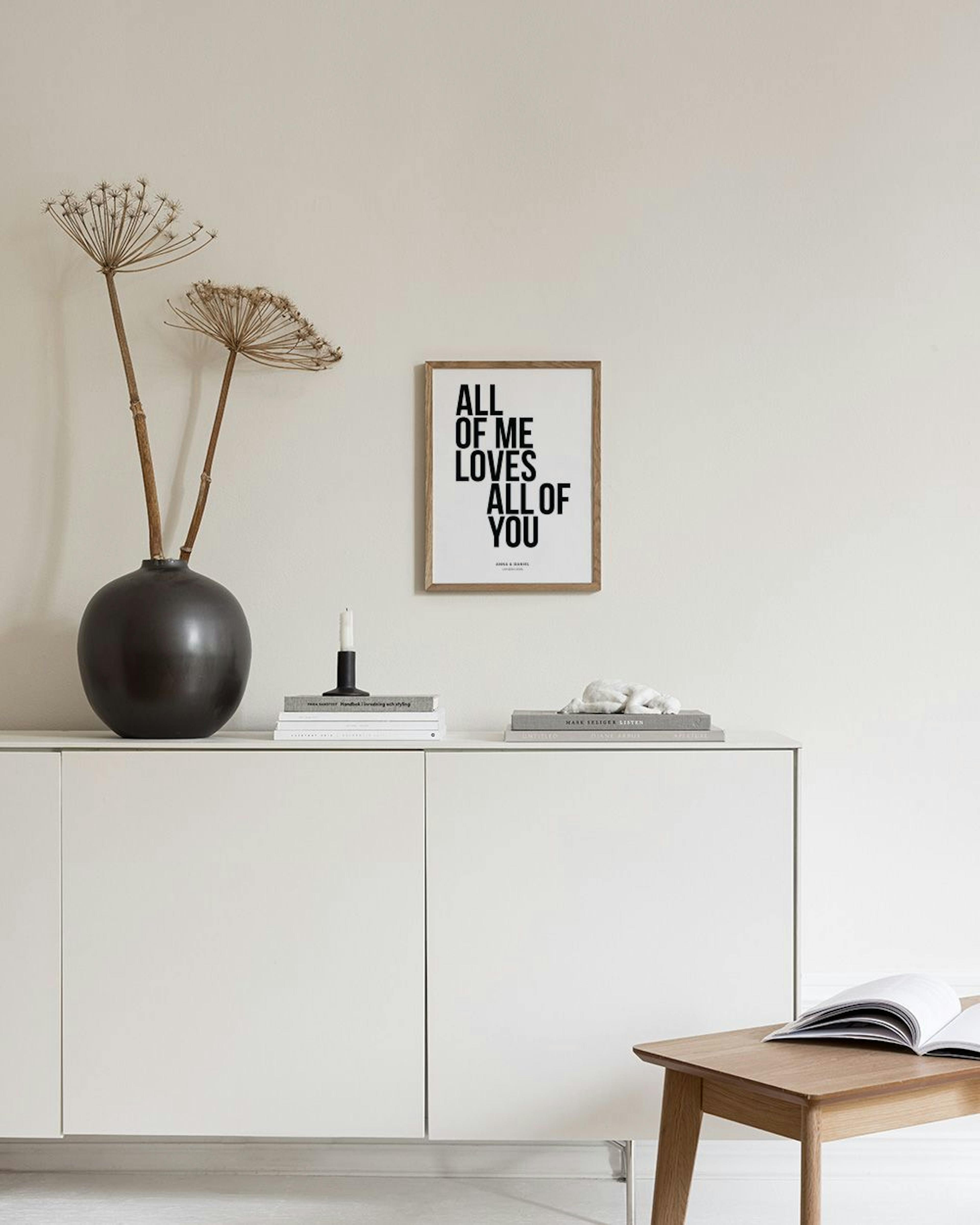 A personalised print with black text All of me loves all of you and names, displayed above a dark shelf with a charcoal vase and