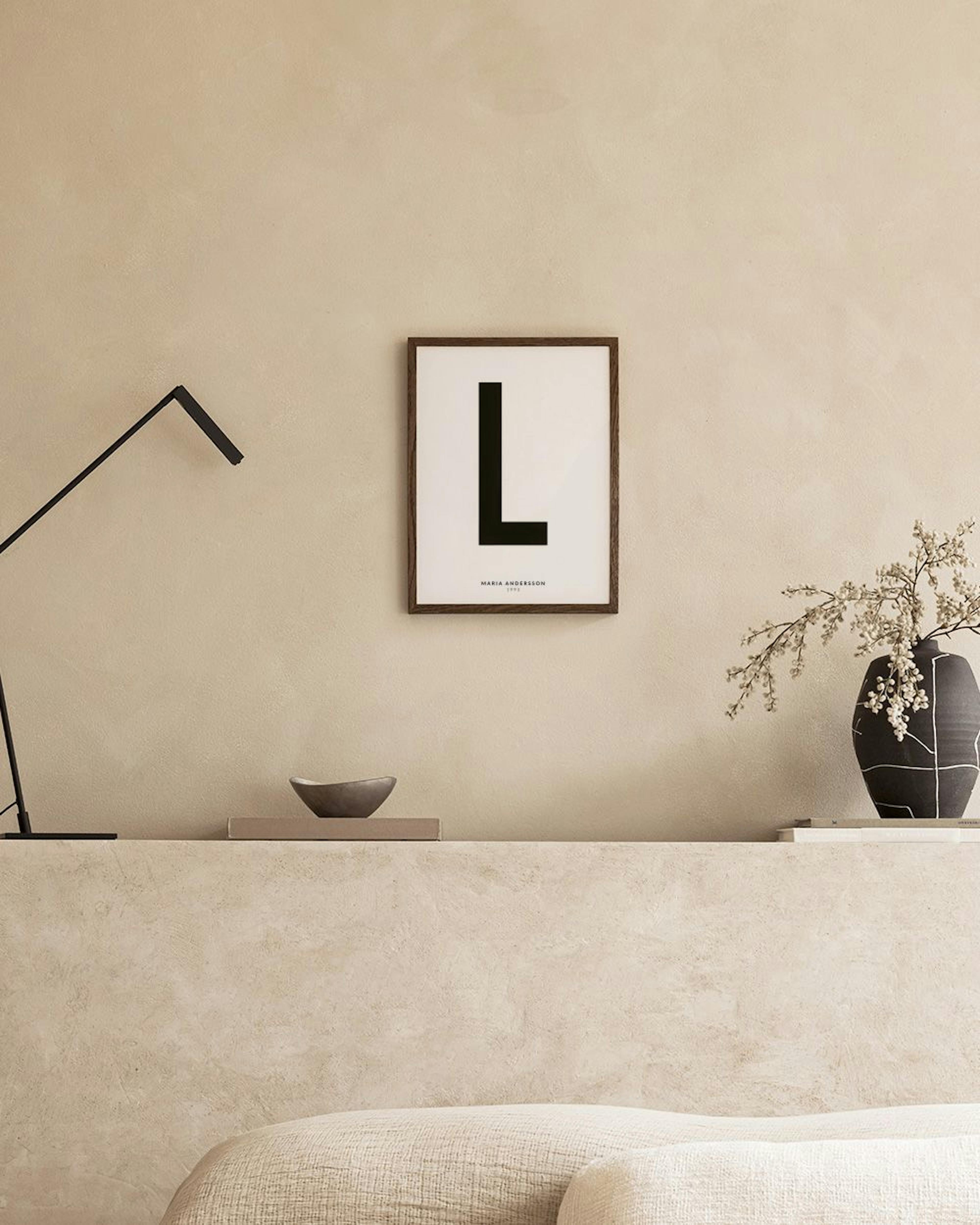 Poster personalizado com a letra L preta em fundo branco, nome e data, numa parede cinzenta, com banco e mesa lateral.