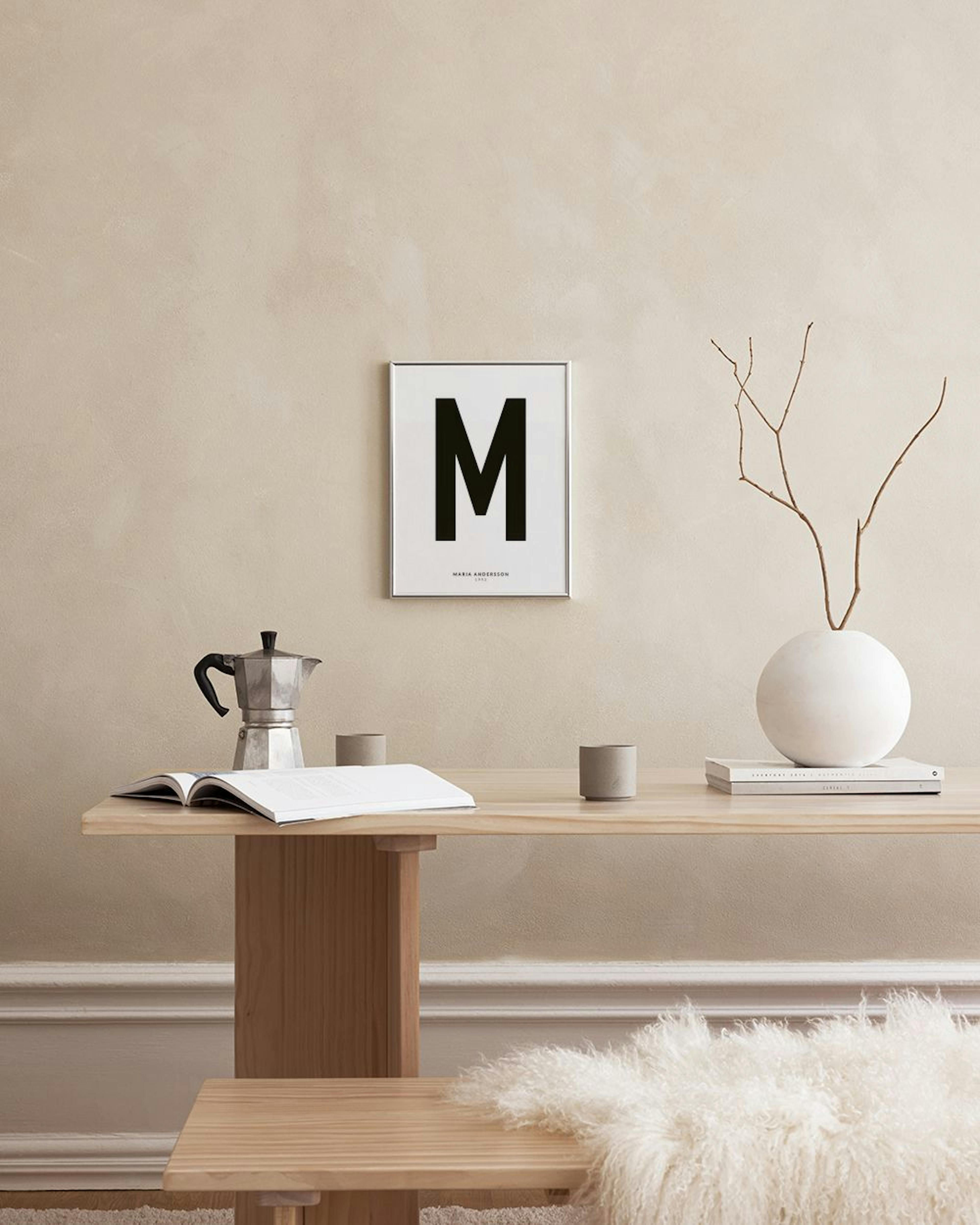 A personalized print with a large black M, MARIA ANDERSSON, and 1993 on a white background, displayed on a grey bench with decor