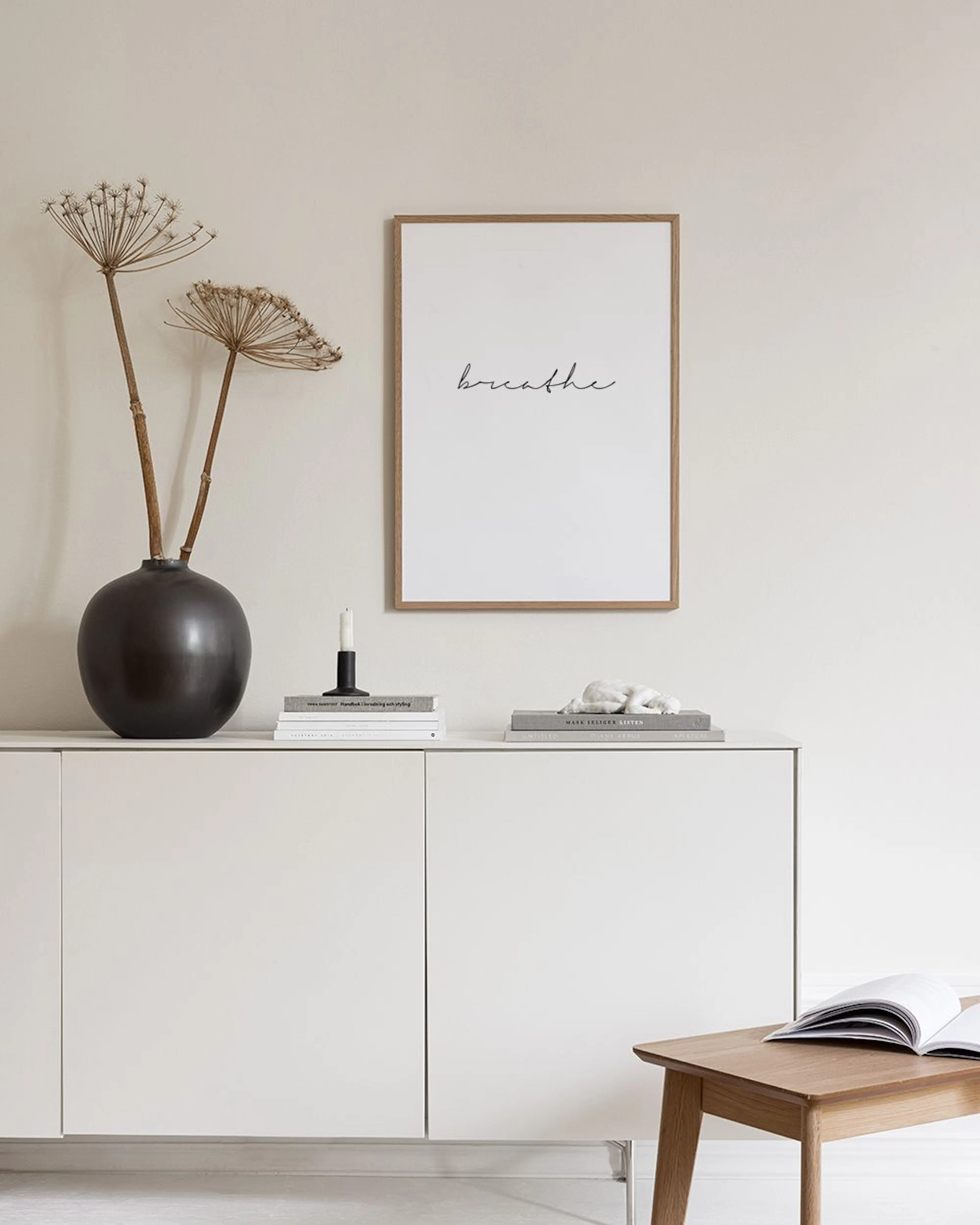 Plakat med ordet breathe i sort skrift, hængende over en træbænk med vaser, bøger og puder i en lys stue.