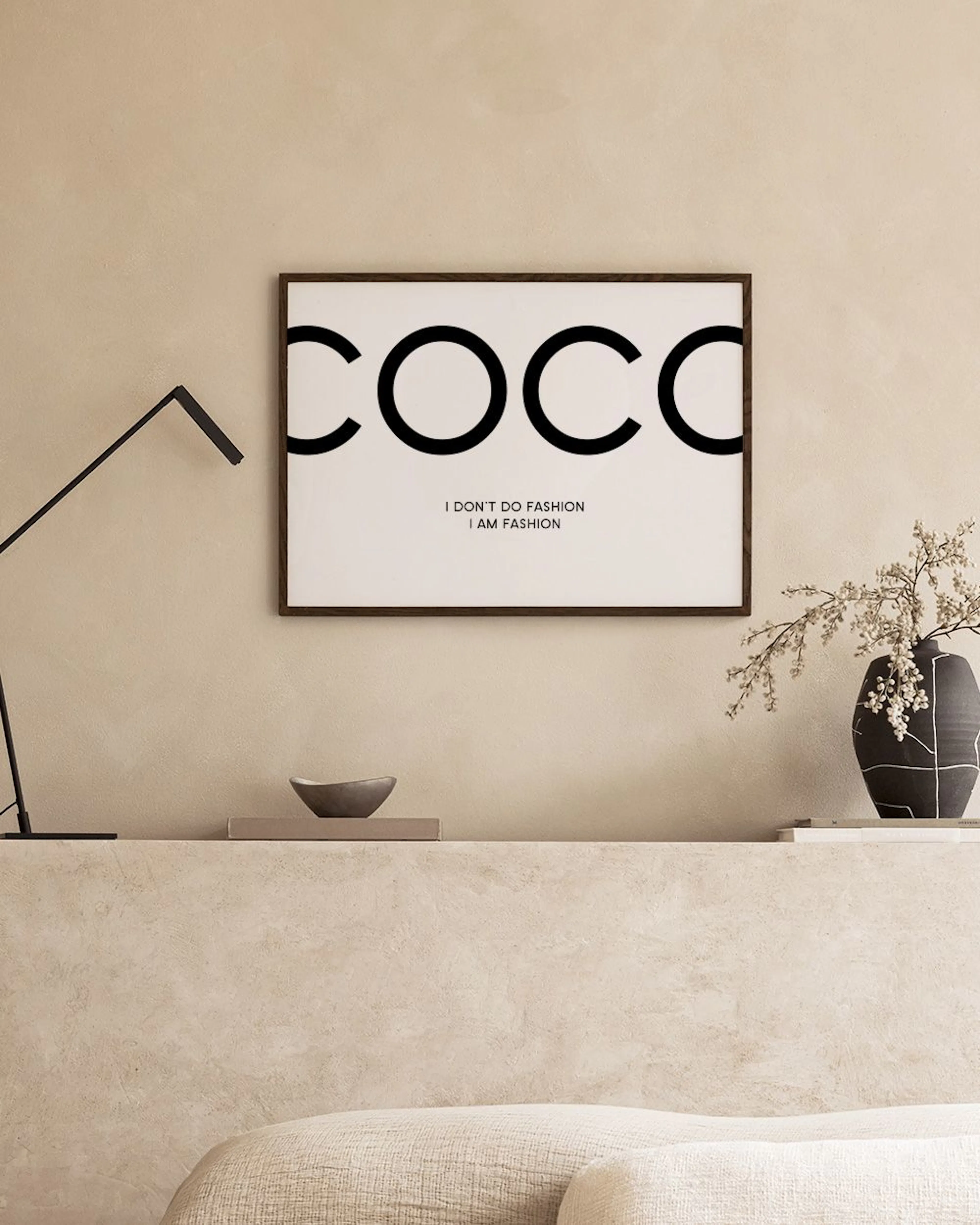 Plakat z napisem COCO, I DONT DO FASHION I AM FASHION, wisi na ciemnoszarej ścianie nad czarnym stolikiem.