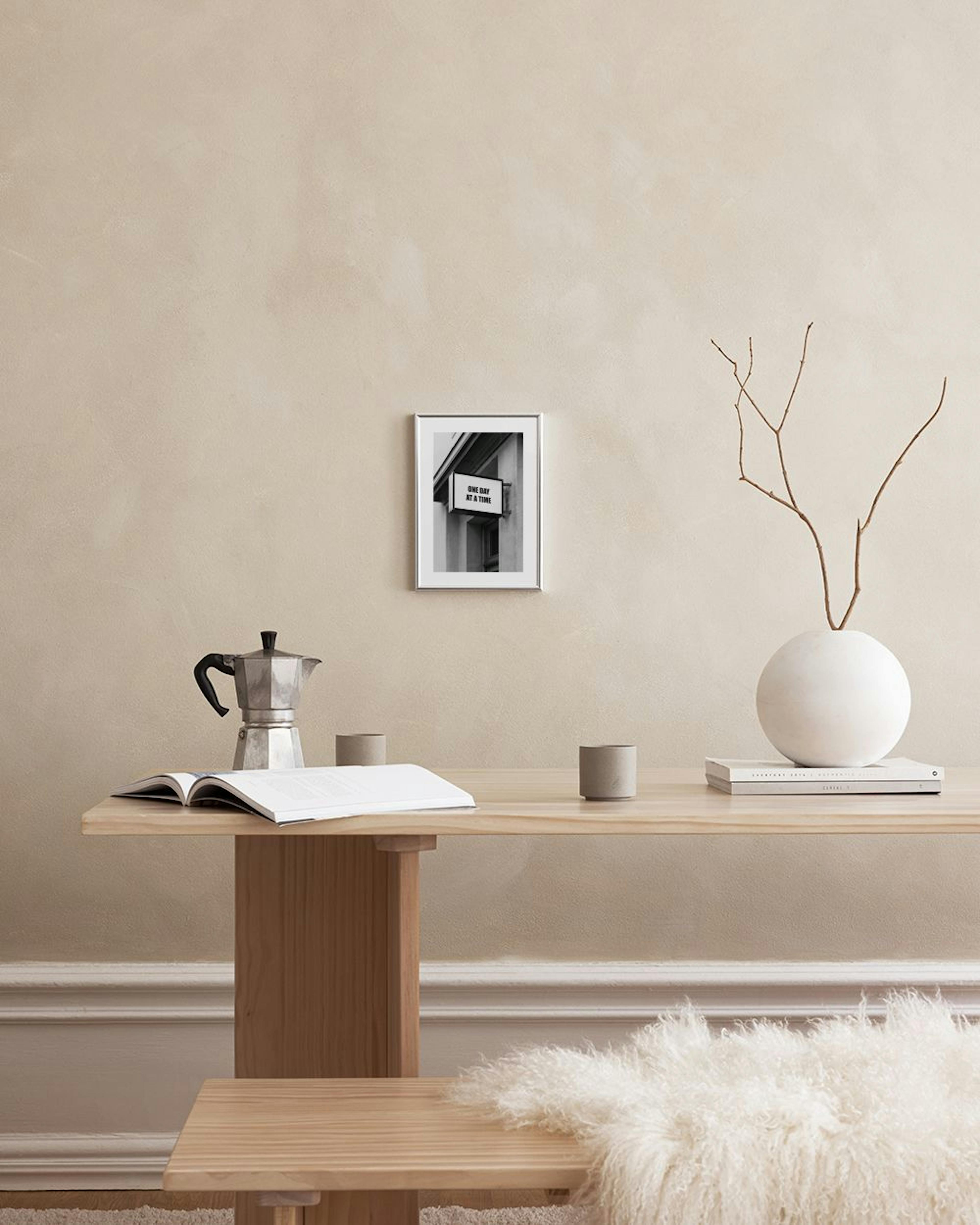 Zwart-wit poster van een uithangbord met de tekst ONE DAY AT A TIME, hangend boven een wit marmeren bijzettafel met vazen.