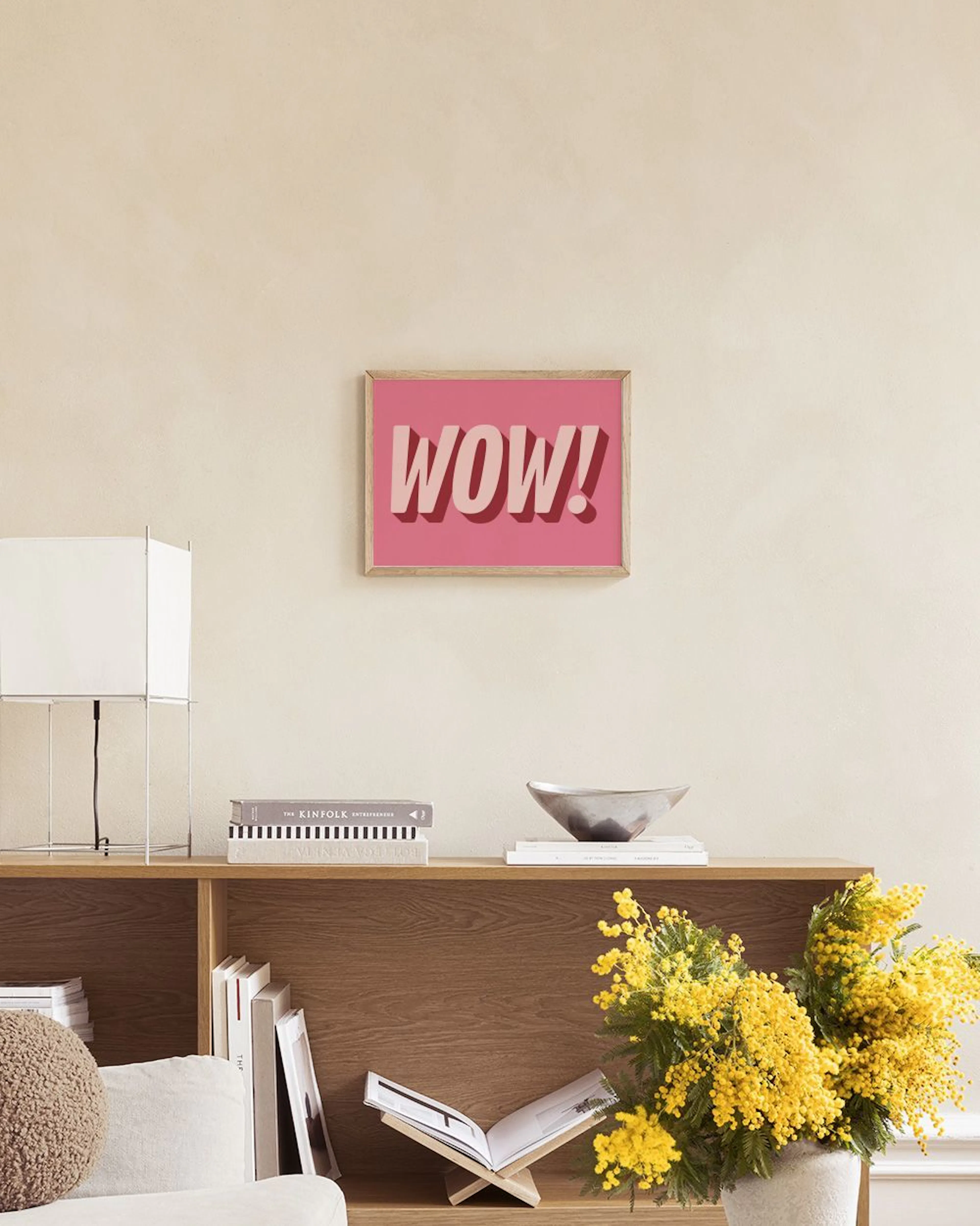 Póster rosa vibrante con la palabra WOW! en letras 3D color rosa claro, exhibido sobre un estante negro con libros y accesorios 