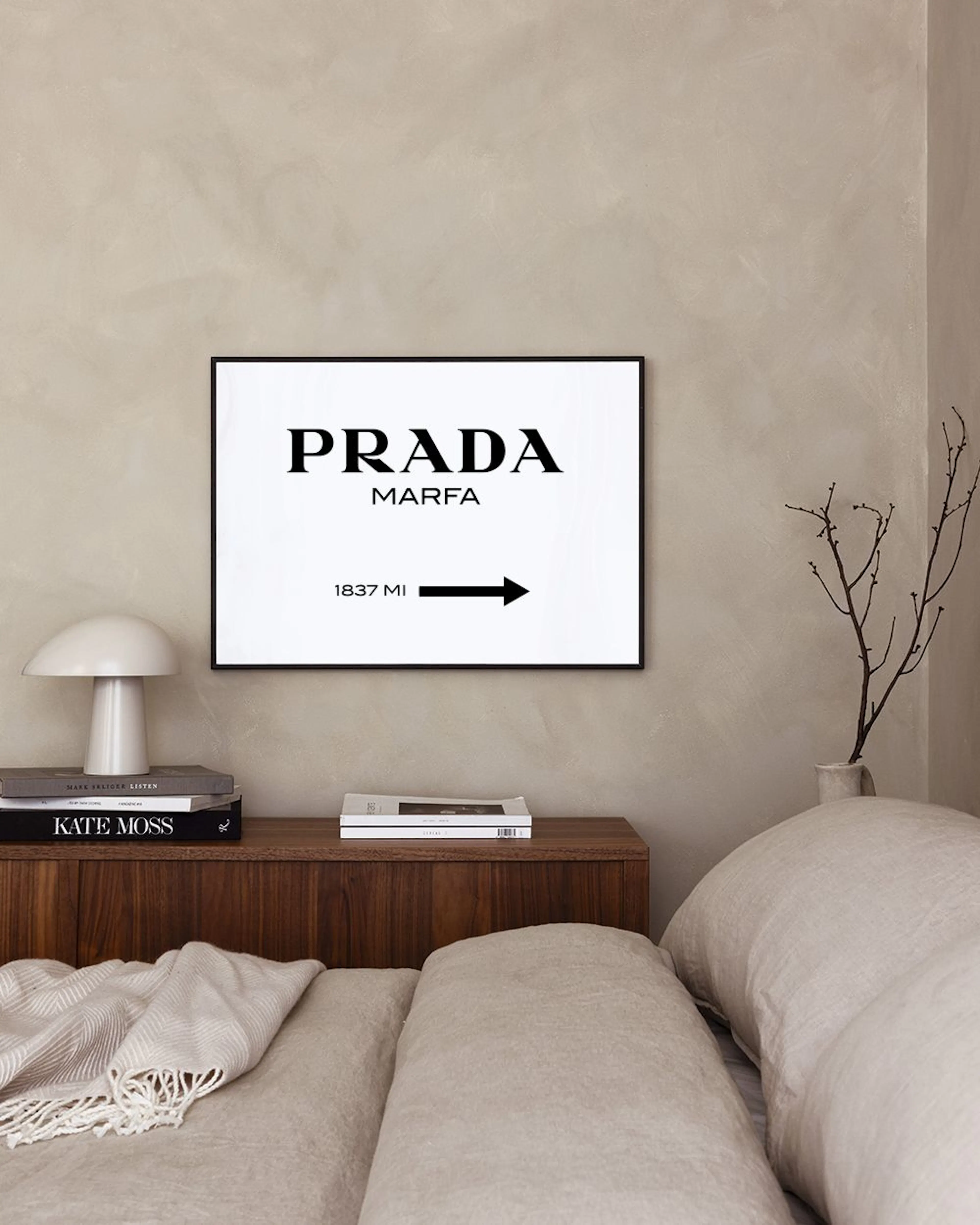 Affiche Prada Marfa noire et blanche, affichée au-dessus dune commode blanche avec lampe et plante.