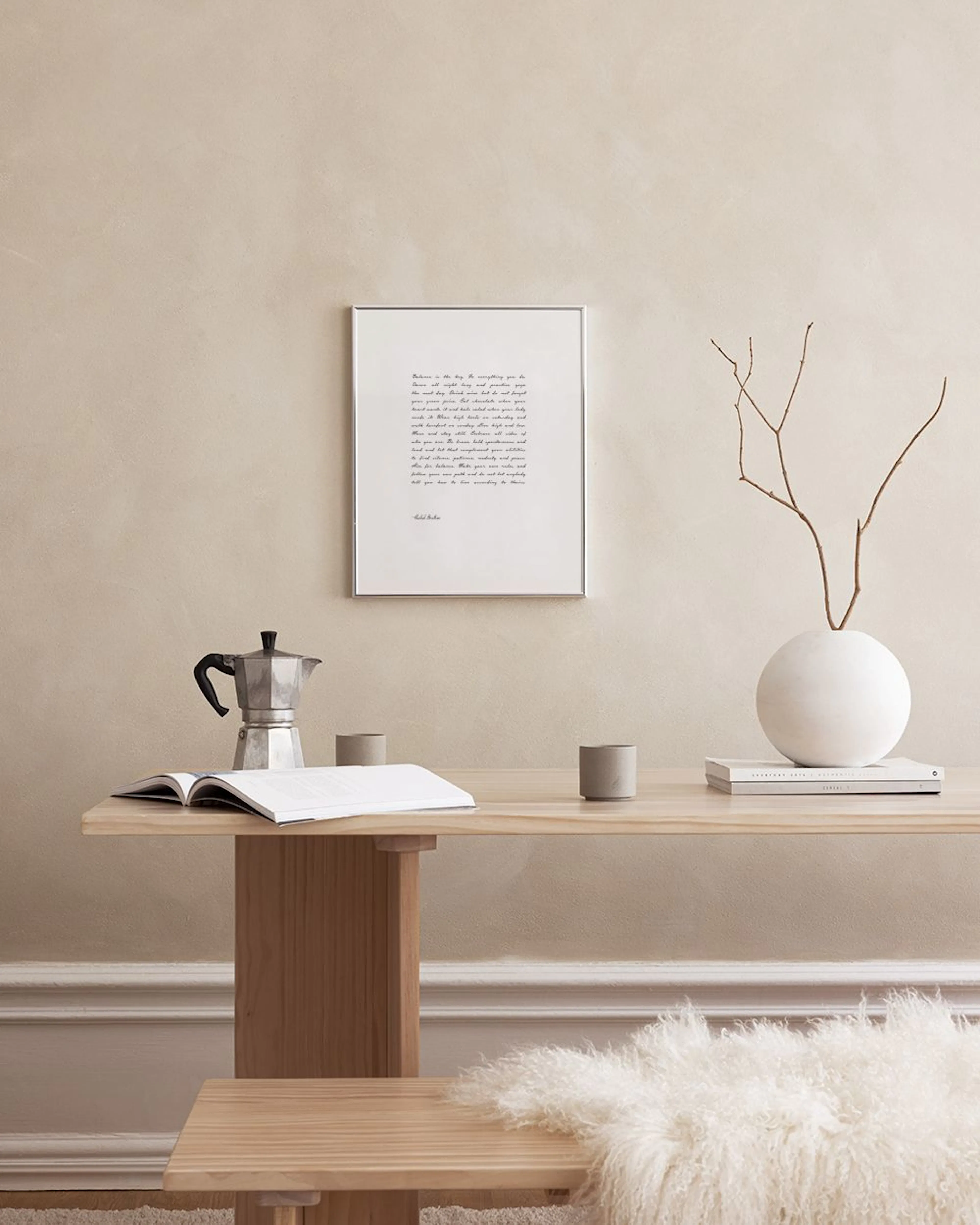 A text print with a handwritten quote in black on a white background, hanging above a wooden bench with a plant and decor.