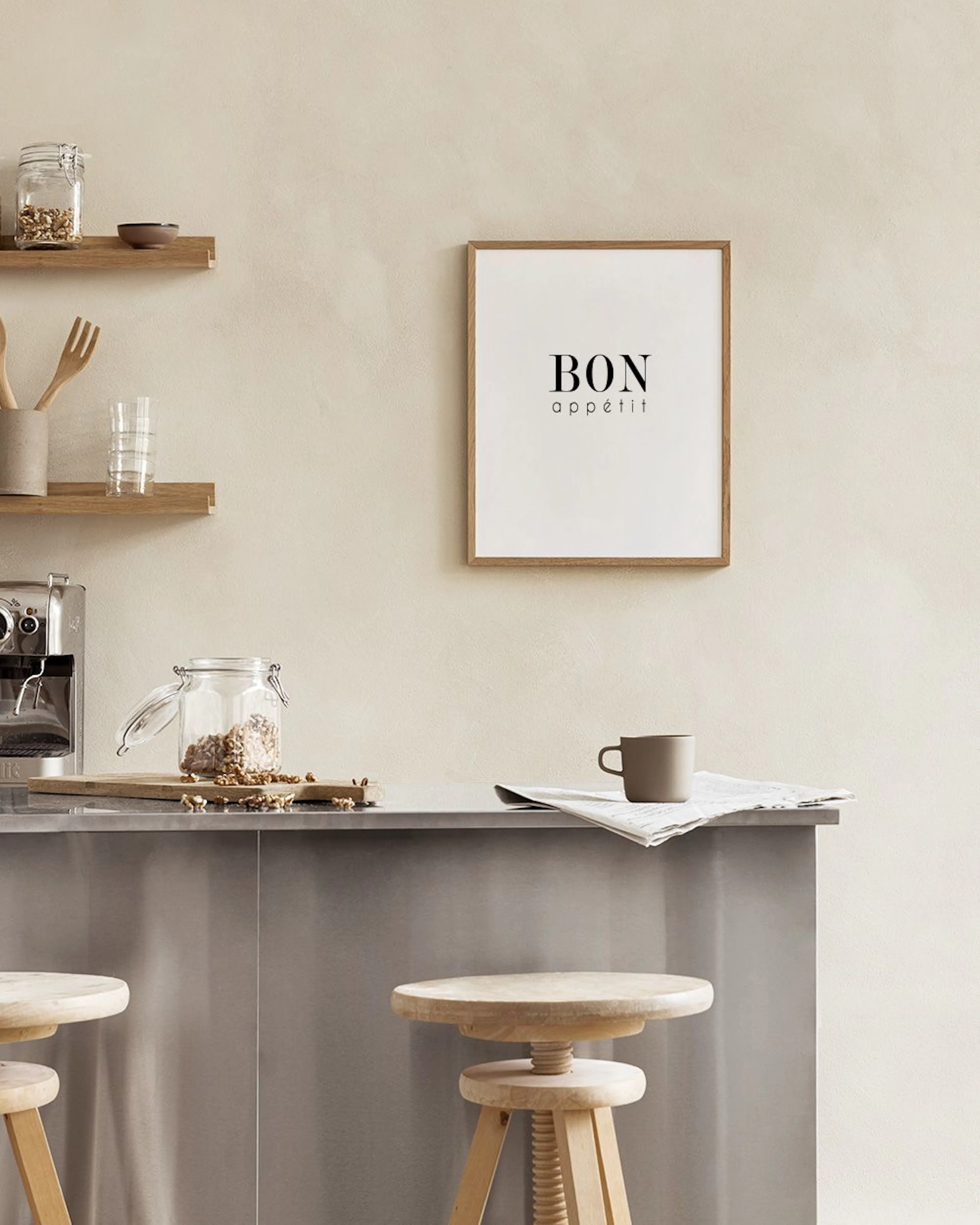 Poster med texten BON appétit i svart, stående på en bänk med marmorbakgrund, bredvid en växt och köksredskap.