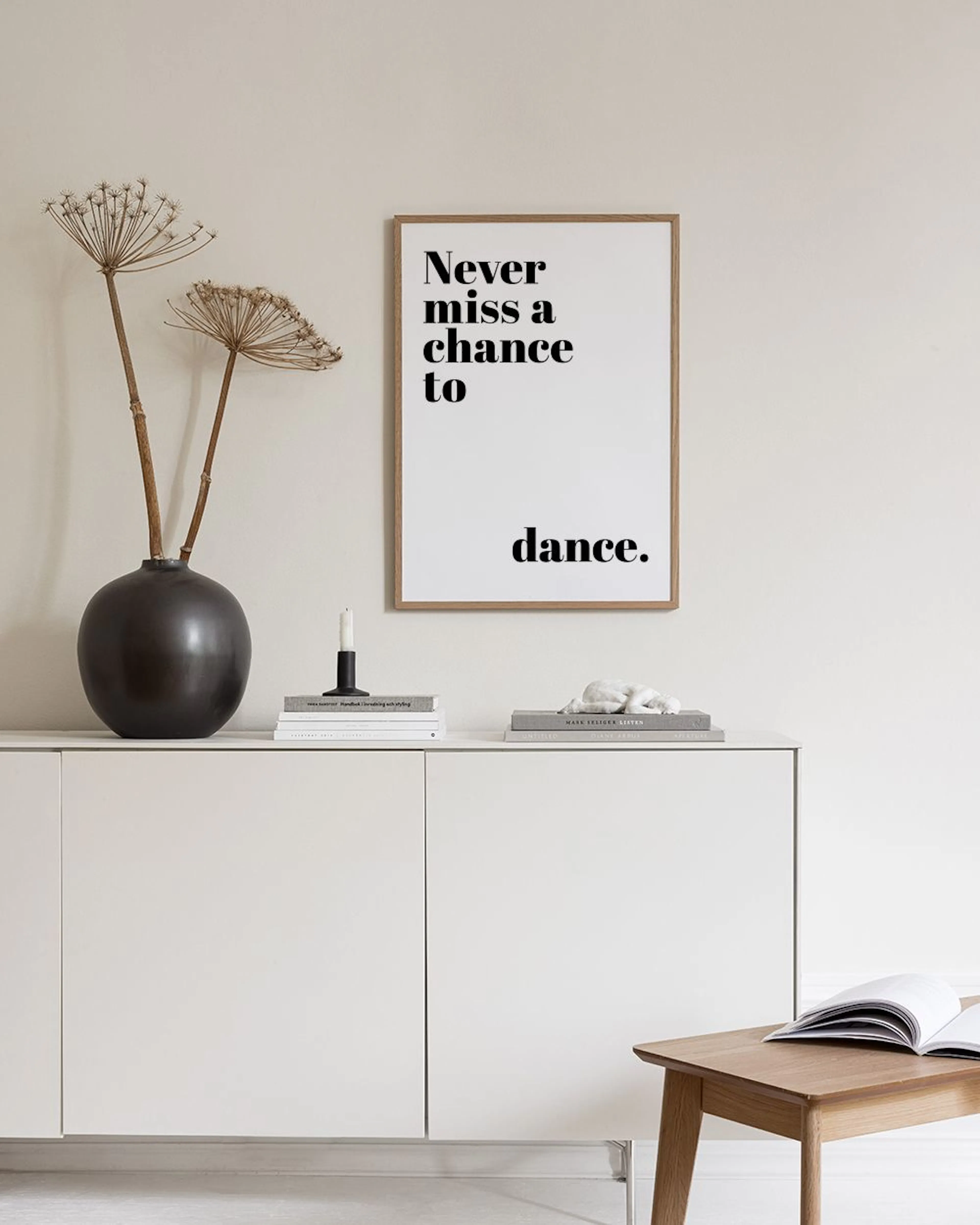 Poster con testo nero Never miss a chance to dance. su sfondo bianco, appeso sopra una credenza bianca con libri e vaso.