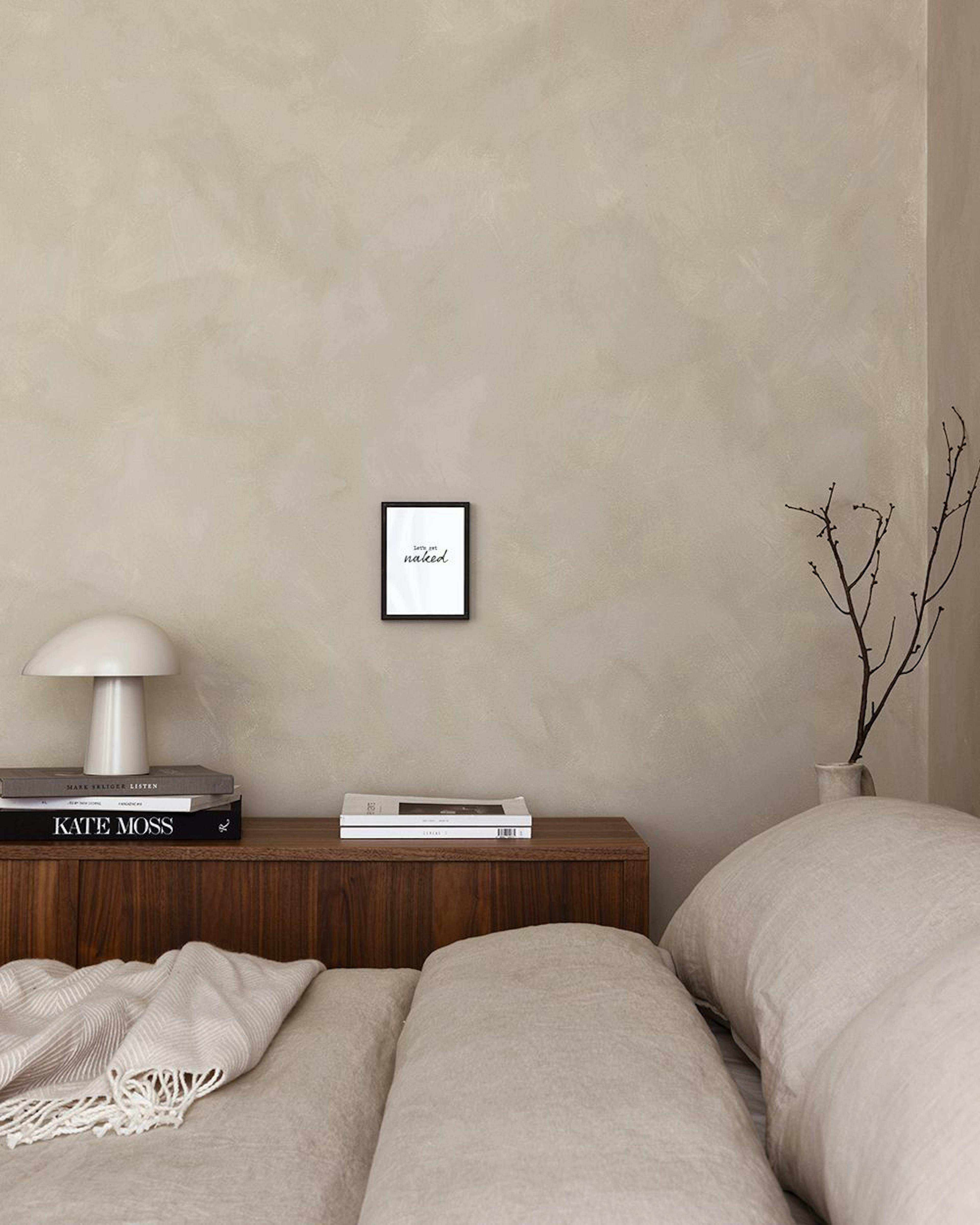 A minimalist print with Lets get naked in black text, hanging on a light grey wall above books and decorative items.
