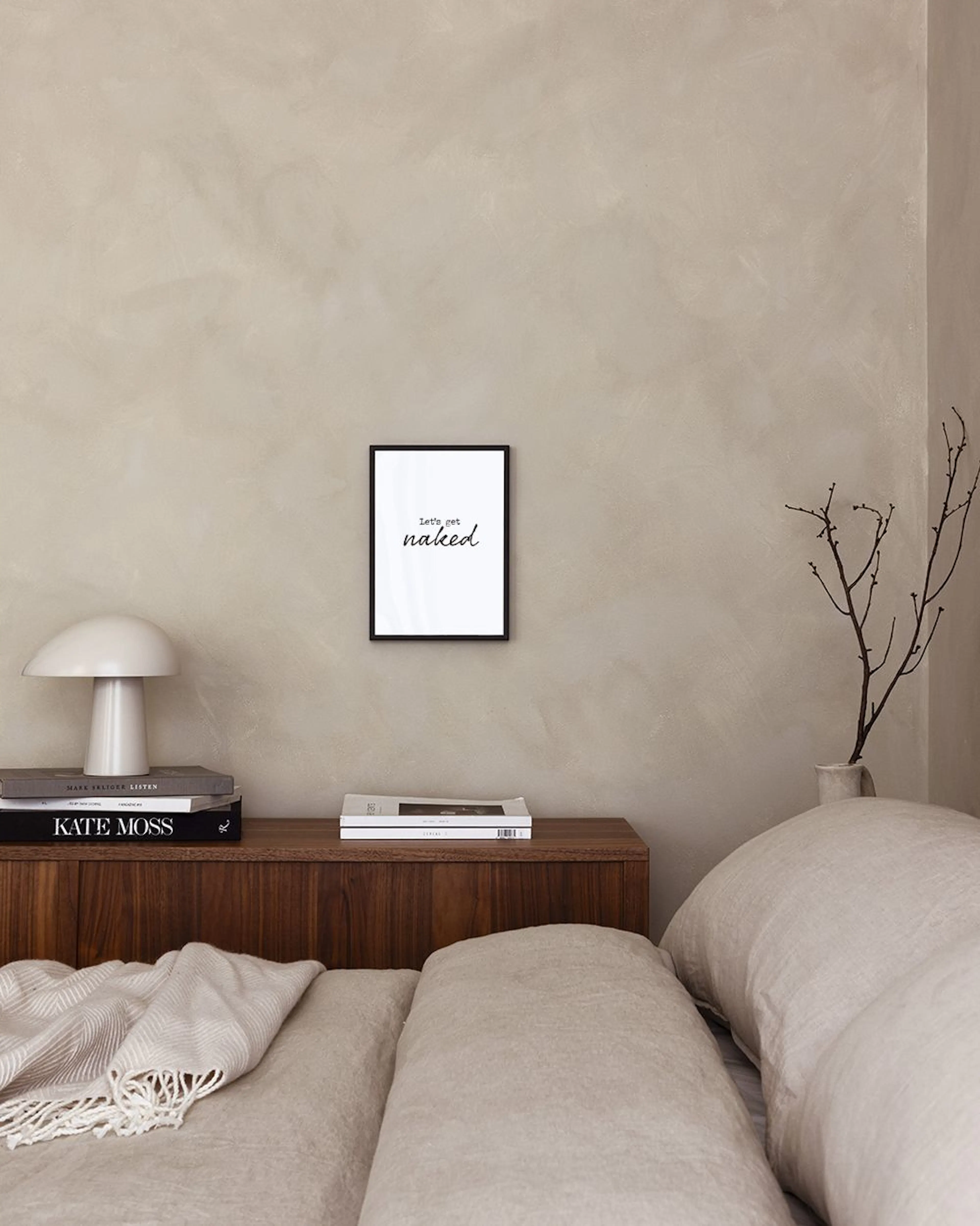 Witte poster met de tekst Lets get naked in zwart, hangend in een kamer boven een stapel boeken en decoratie.