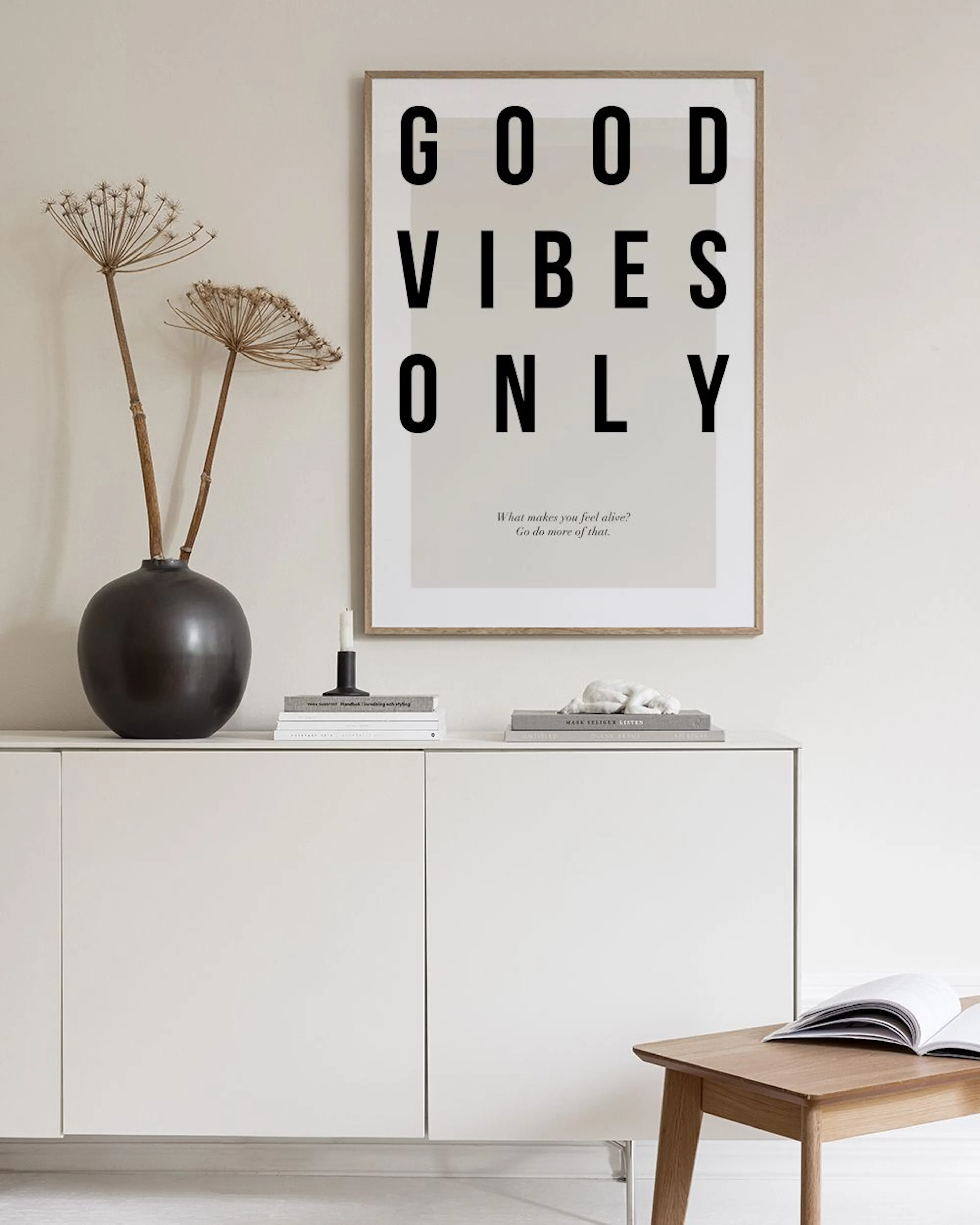 Póster com GOOD VIBES ONLY em preto sobre fundo bege, pendurado numa parede cinzenta, com um vaso e cadeira.