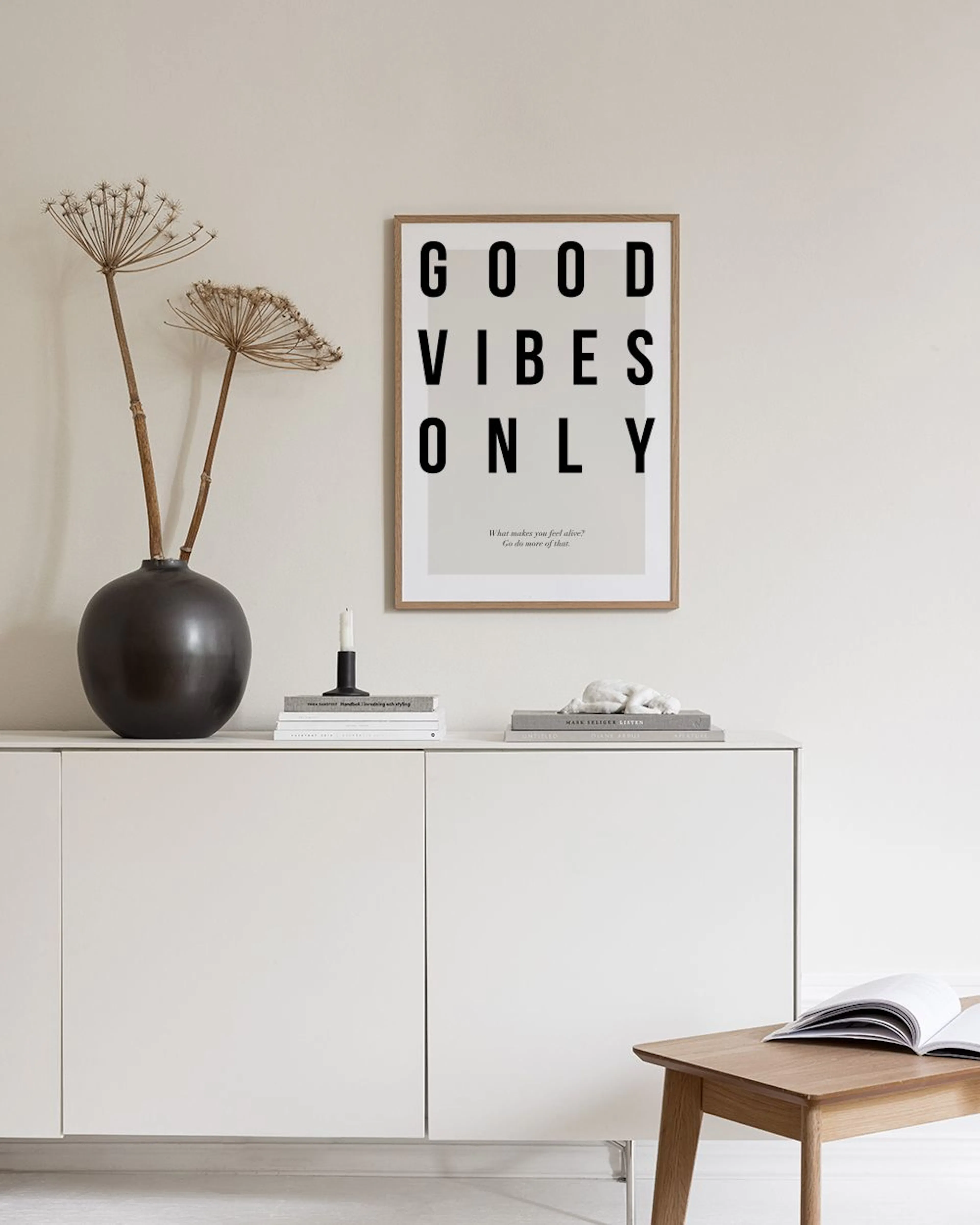 A Good Vibes Only typography print on a gray wall with a white vase, books, and a cane chair.
