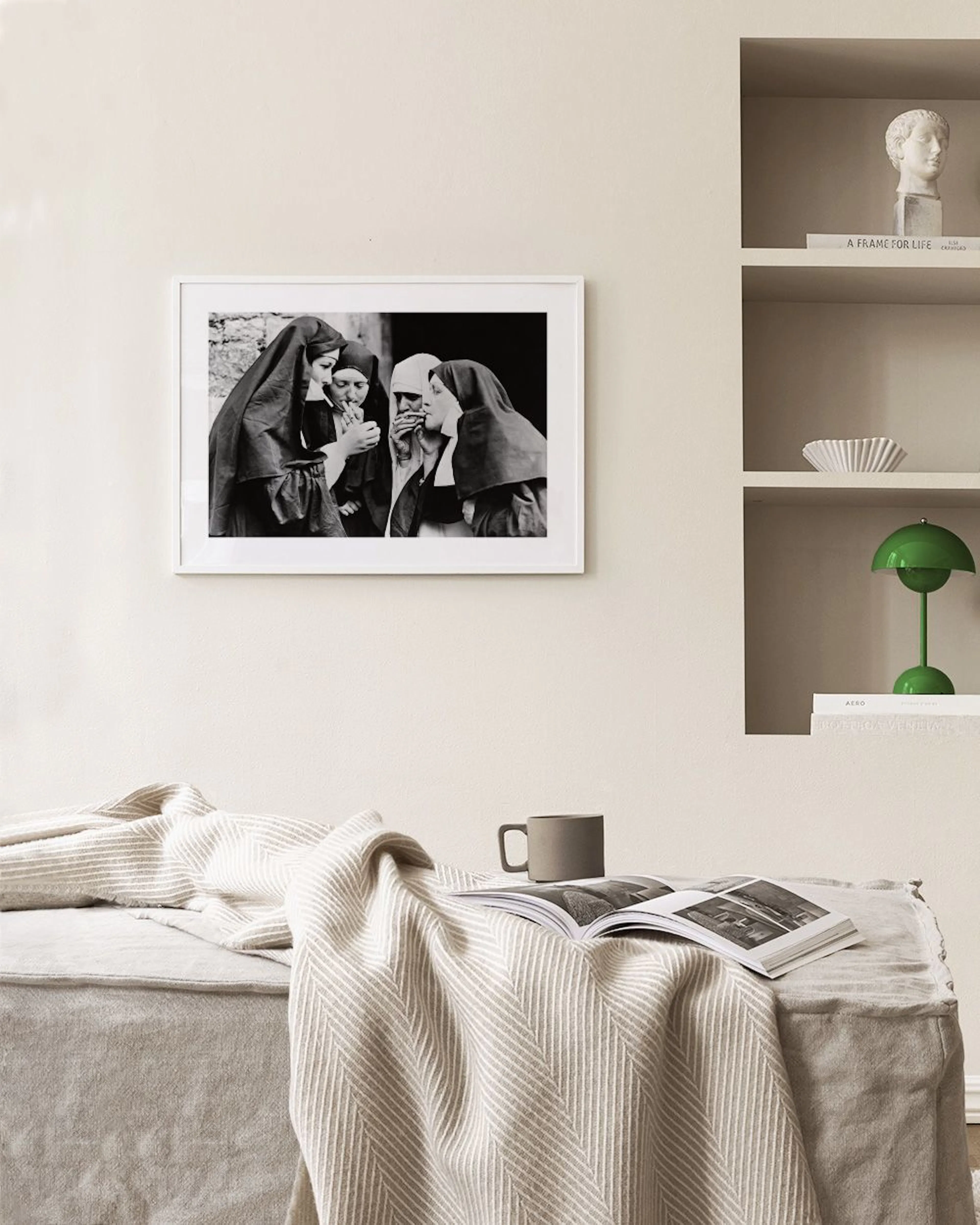 Affiche en noir et blanc de nonnes allumant une cigarette, accrochée au-dessus dune table basse grise et dune chaise en velours.