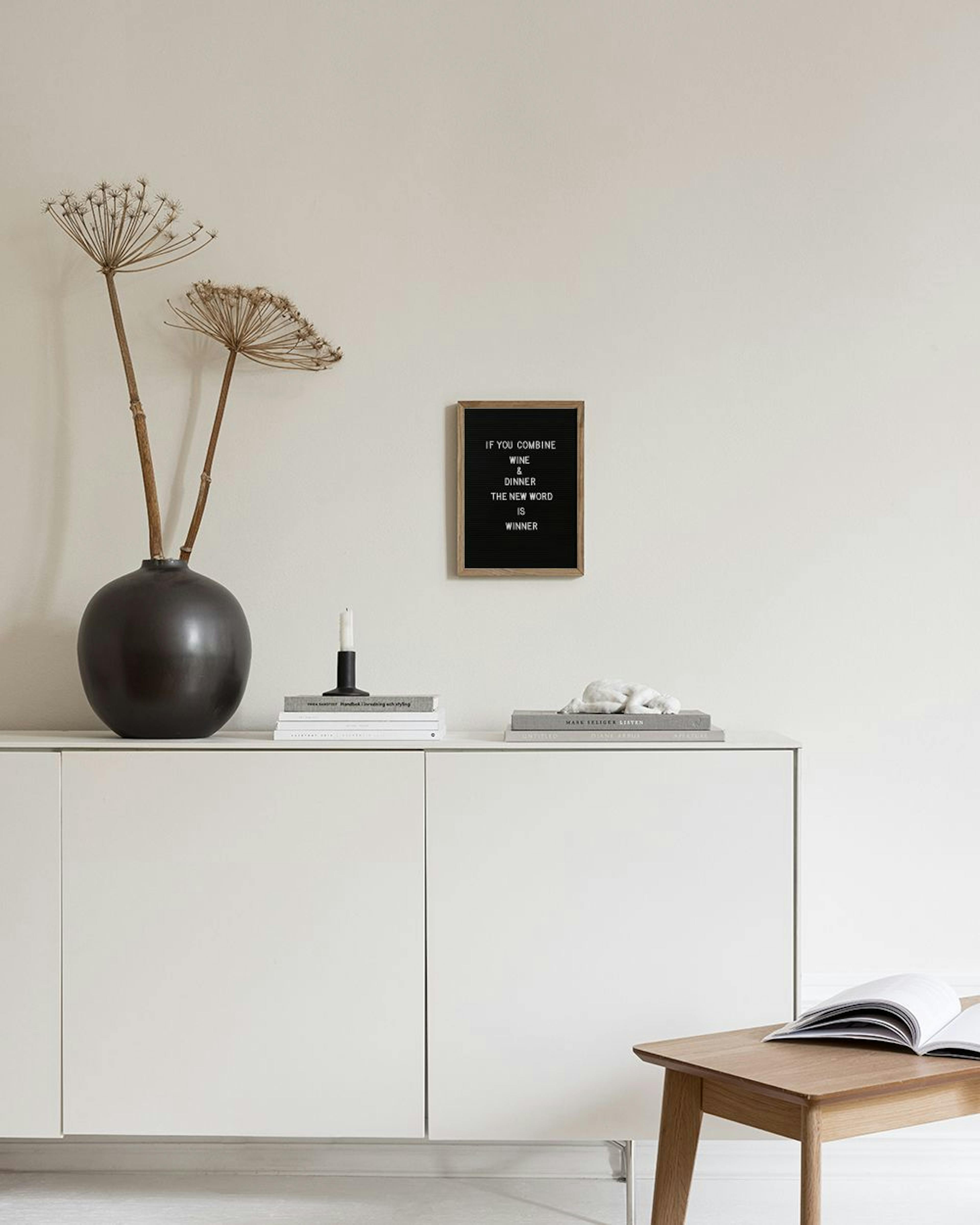 A black letter board print with a quote about wine and dinner, hanging above a white dining table with a book and mug.