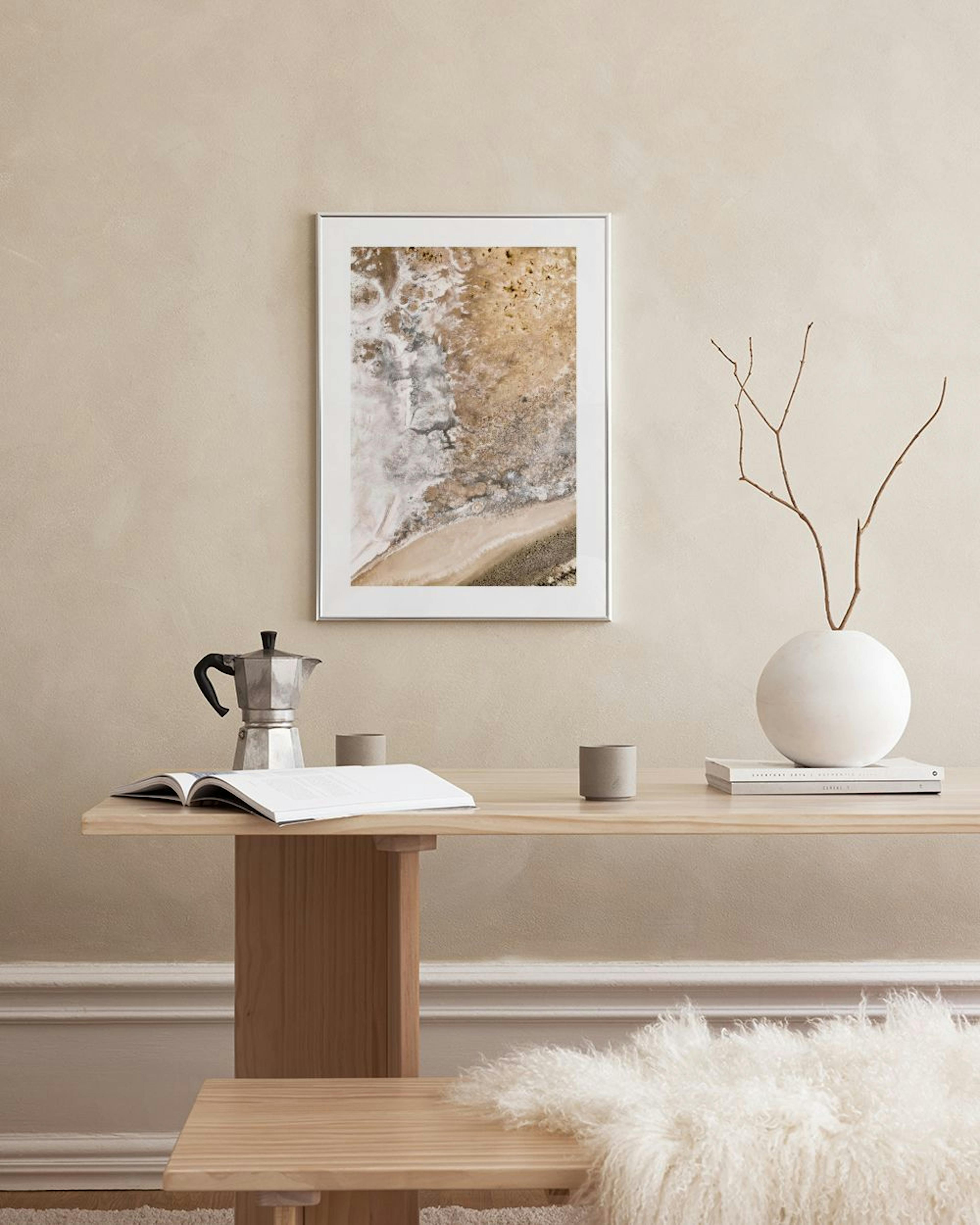 A print of a rocky beach with sand, water, and pebbles in beige, white and brown tones, hanging above a wooden chair.