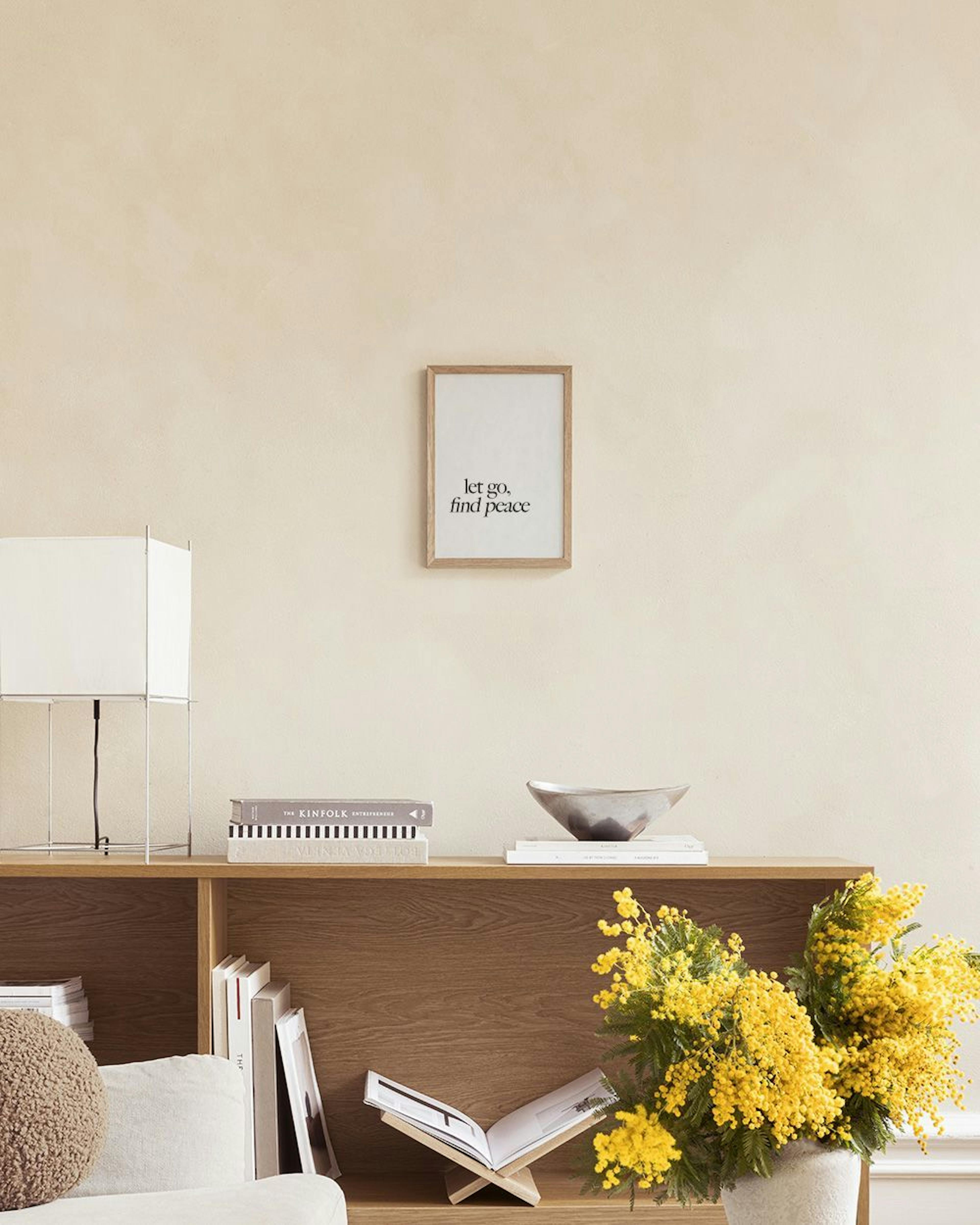 A minimalist print with let go, find peace in black script, hanging above a shelf with an open book, stacked pebbles, and a meta