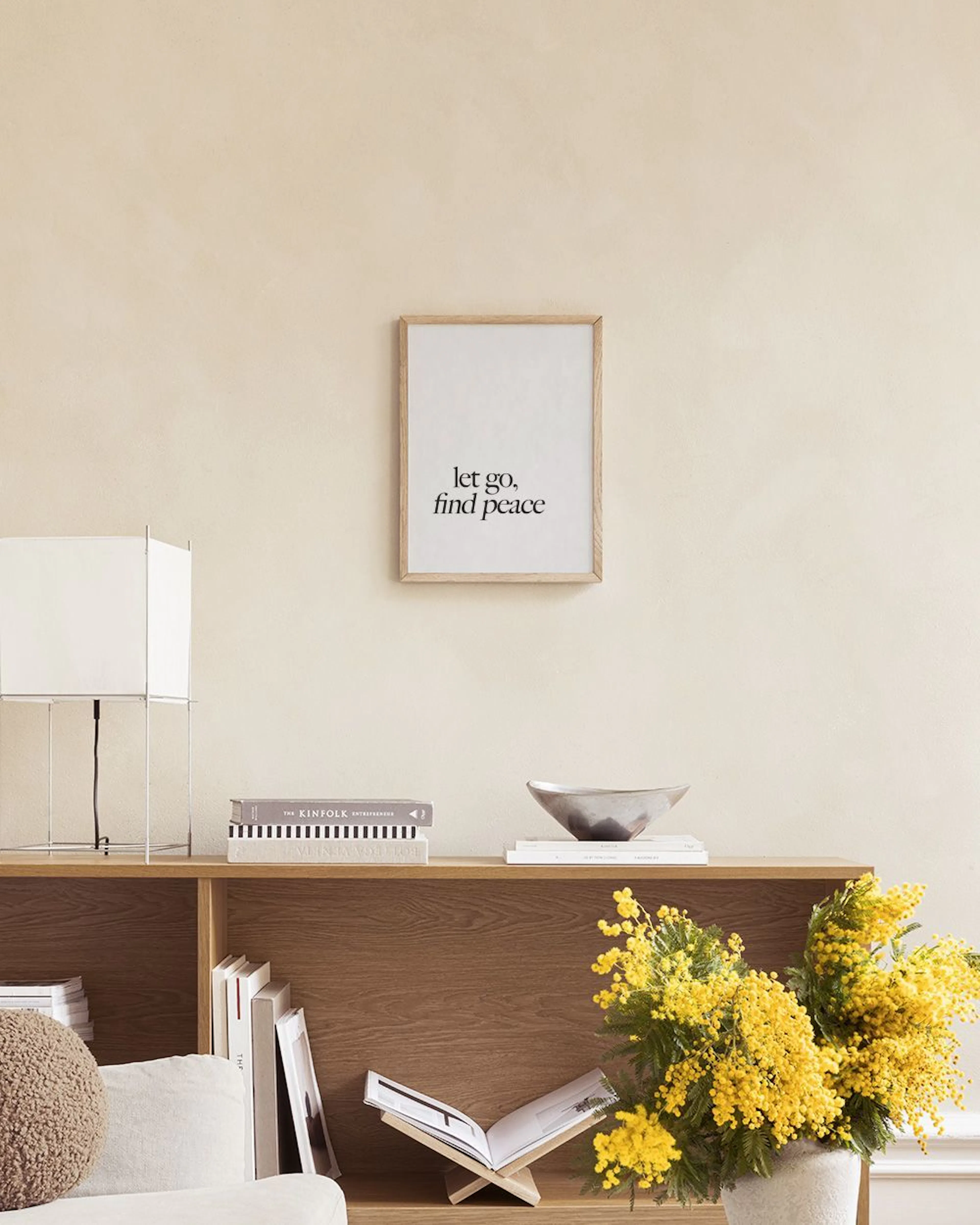 A minimalist print with the black text let go, find peace hanging above a white shelf with an open book, stacked pebbles, and a 