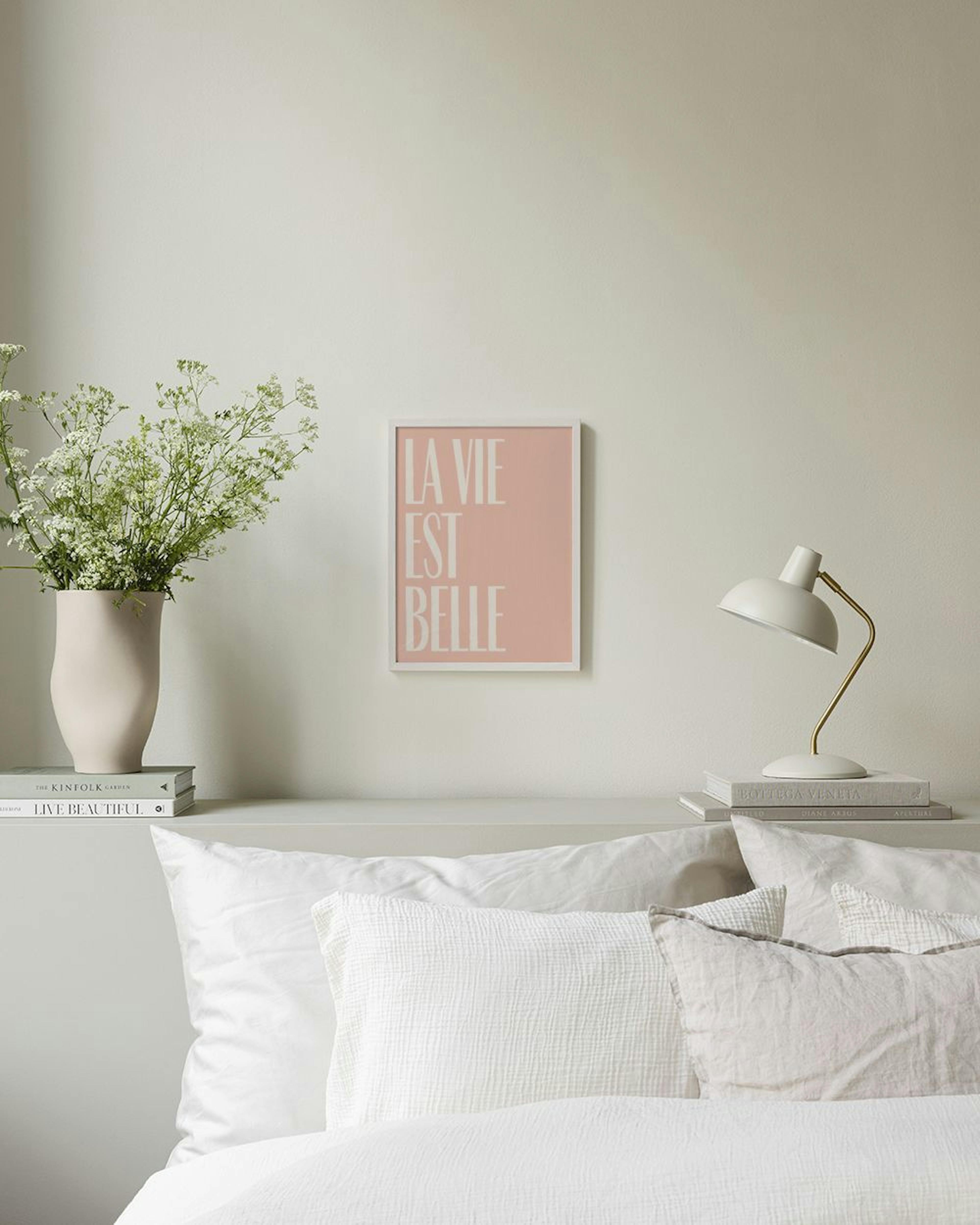 Póster rosa de La Vie Est Belle con letras blancas, colgado sobre un mueble de madera y ratán, con decoración boho.