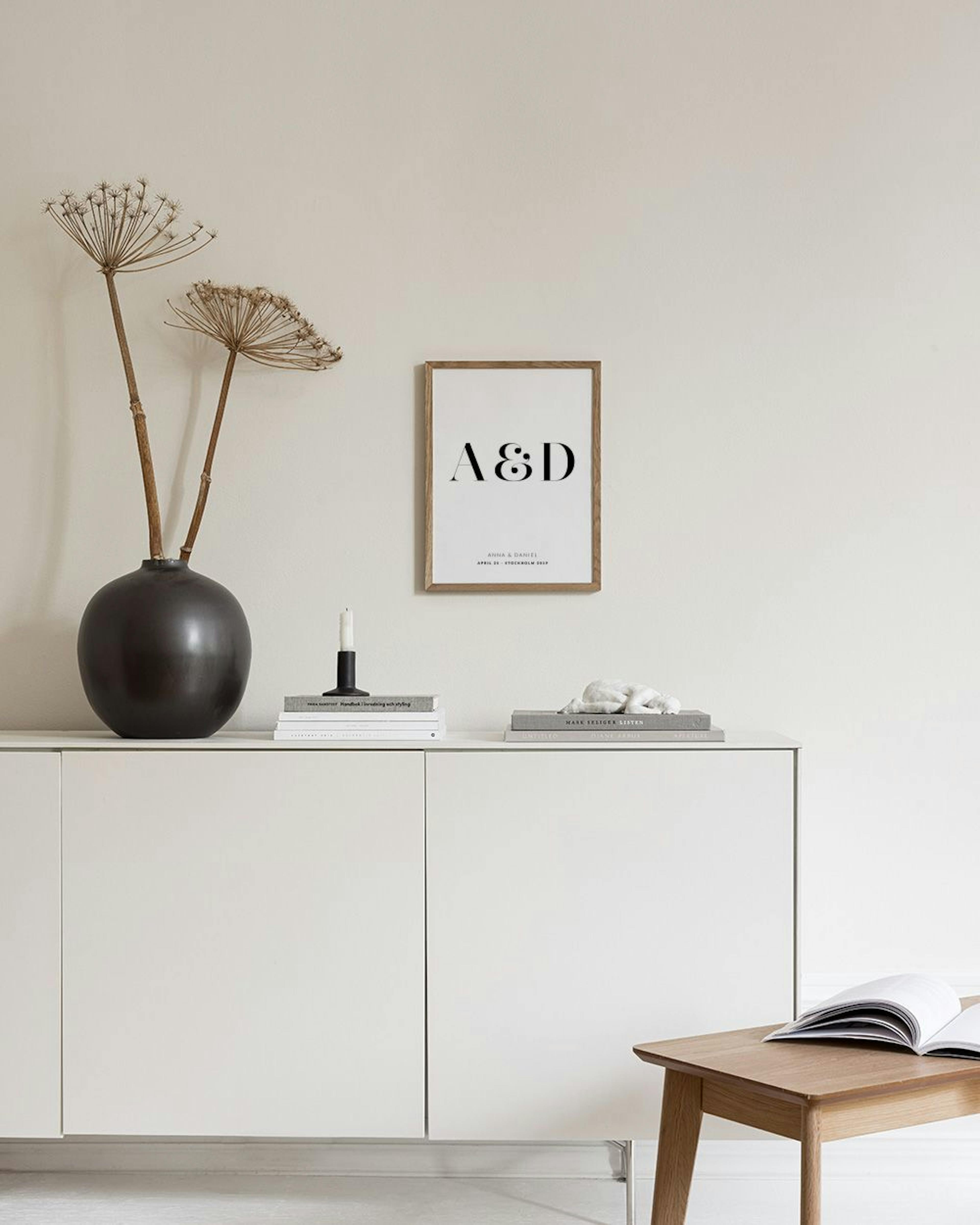 Póster personalizado con las iniciales A&D en negro, nombres y fecha, colgado en un dormitorio sobre una mesa de noche.