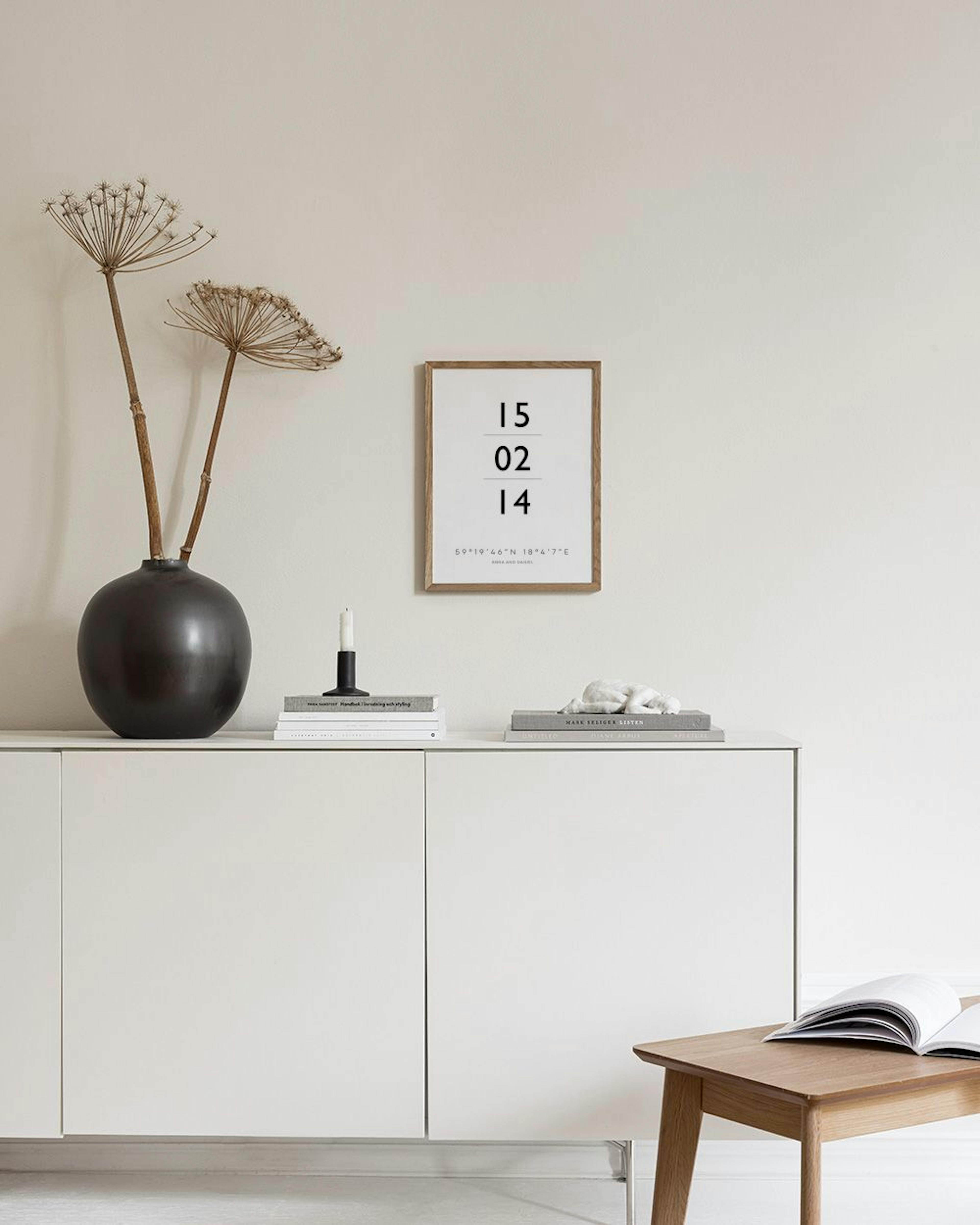 Póster personalizado blanco con números negros y coordenadas, colgando sobre una banca de madera y mimbre con jarrones y libros.